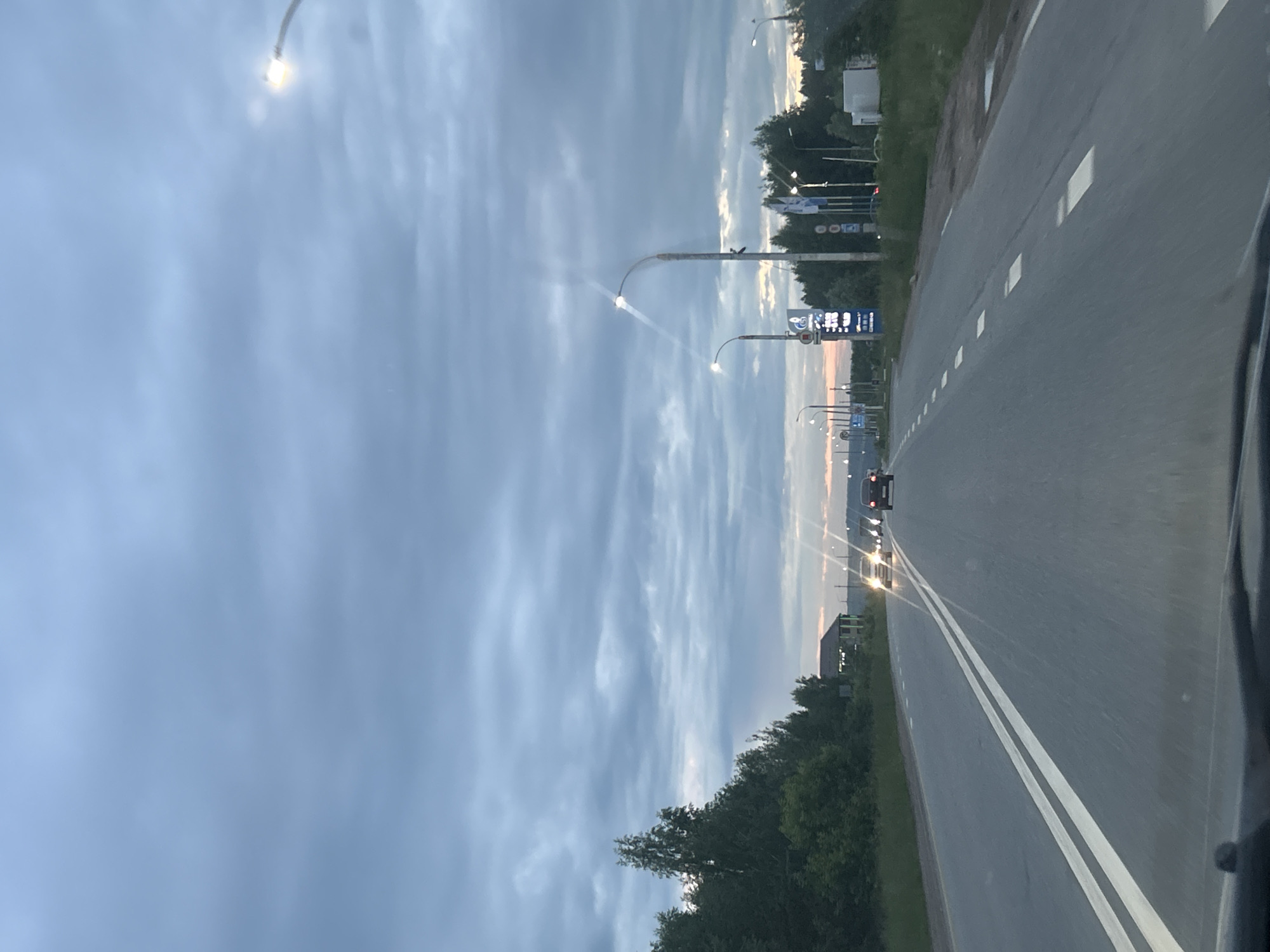 The image shows a perspective from inside a vehicle, looking forward along a two-lane asphalt road. The sky is cloudy and appears to be twilight. On the left side of the road, there are trees and bushes. On the right side of the road, there are streetlights, a gas station with a price sign, and some buildings in the distance. Several cars are visible on the road, some with headlights on. The road has double solid white lines marking the center. The scene is likely on the outskirts of a populated area, given the presence of a gas station and the open landscape beyond the road.