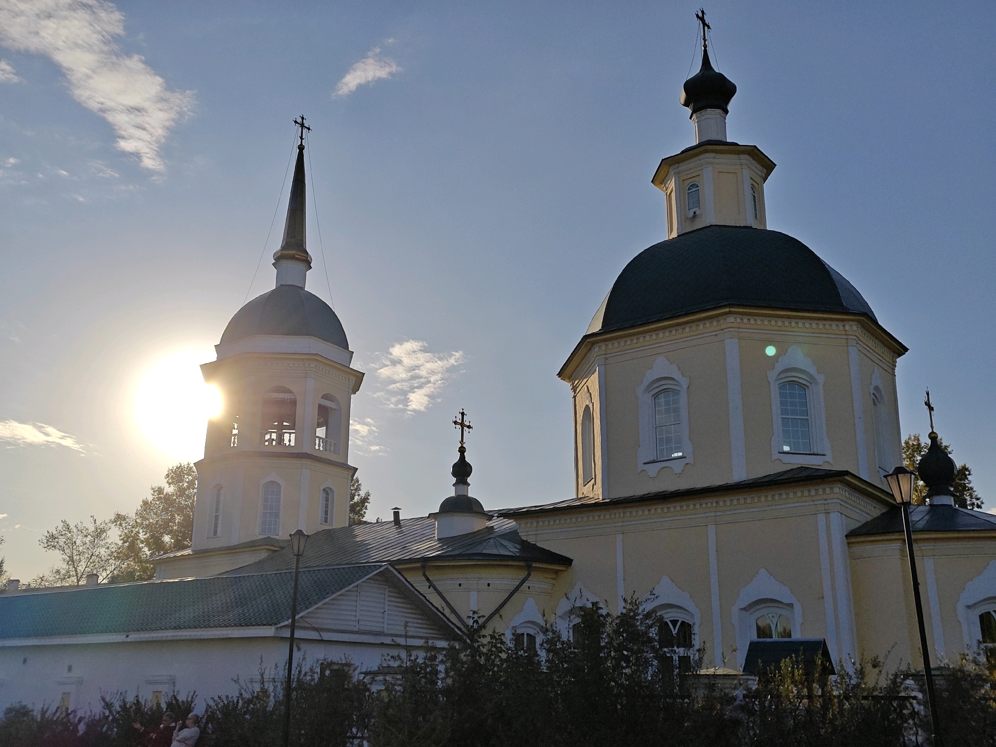 An exterior low-angle shot captures a yellow Orthodox church with a dark green dome, silhouetted against a bright sky with scattered clouds. The sun is partially obscured by a tall bell tower on the left, creating a lens flare effect. The bell tower is topped with a spire and a cross. A larger, domed structure with a cross also rises from the main body of the church. Trees and foliage partially obscure the lower portion of the building, and two individuals are visible standing near the bushes in the foreground. A lamppost stands to the right of the main church structure. The image is taken in Irkutsk, Russia, a city in Eastern Siberia known for its numerous Orthodox churches.