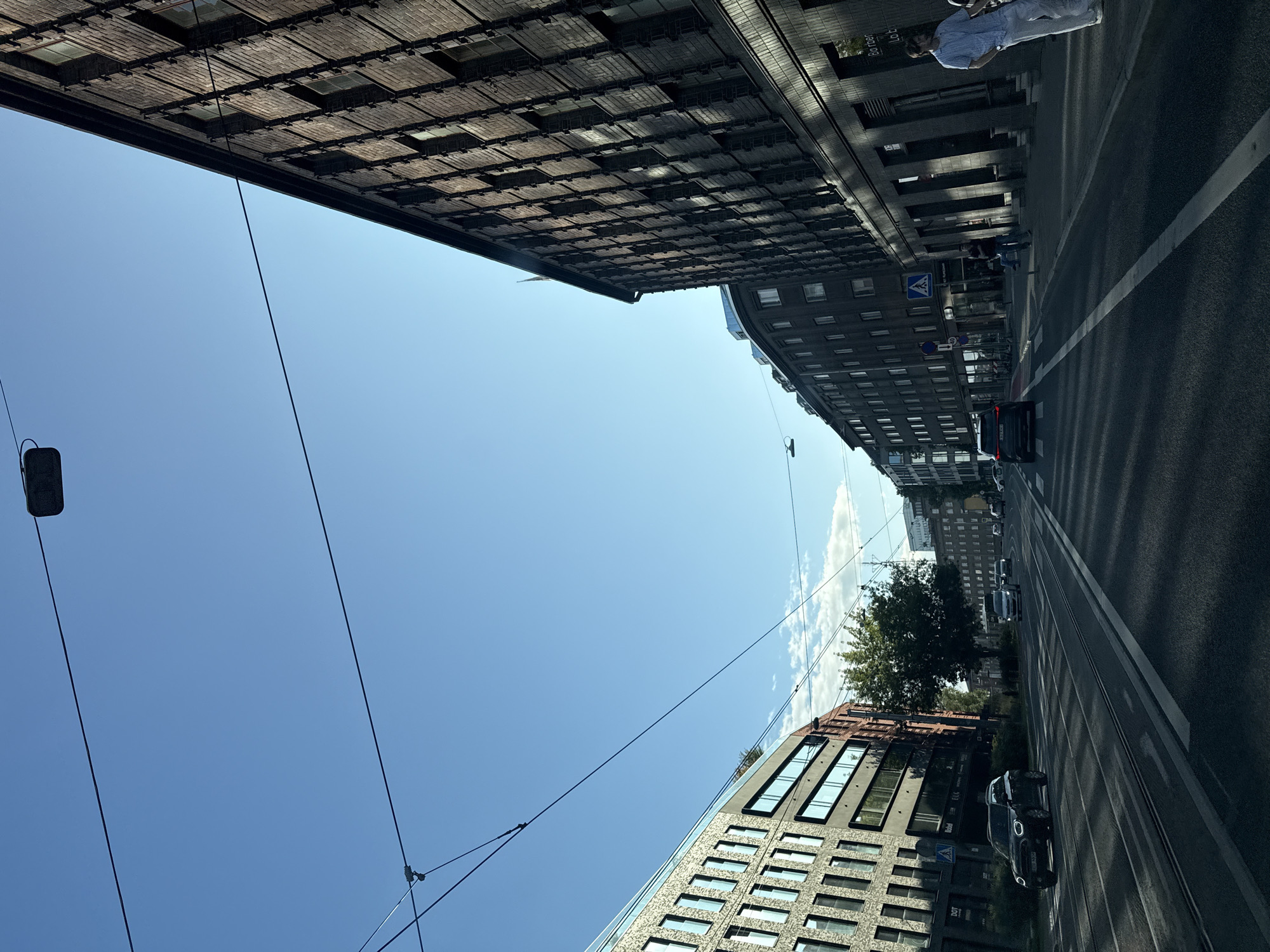 A street view in Tallinn, Estonia shows a street with tram lines and cars. On the right is a dark brick building with many windows. On the left is a modern building with a facade of many windows. Both buildings are multi-story. Several cars are driving on the street. A few pedestrians are visible on the sidewalk. The sky is clear blue with some scattered clouds. Overhead wires are present, likely for trams or trolleybuses. The lighting suggests it is daytime, with shadows cast across the street.