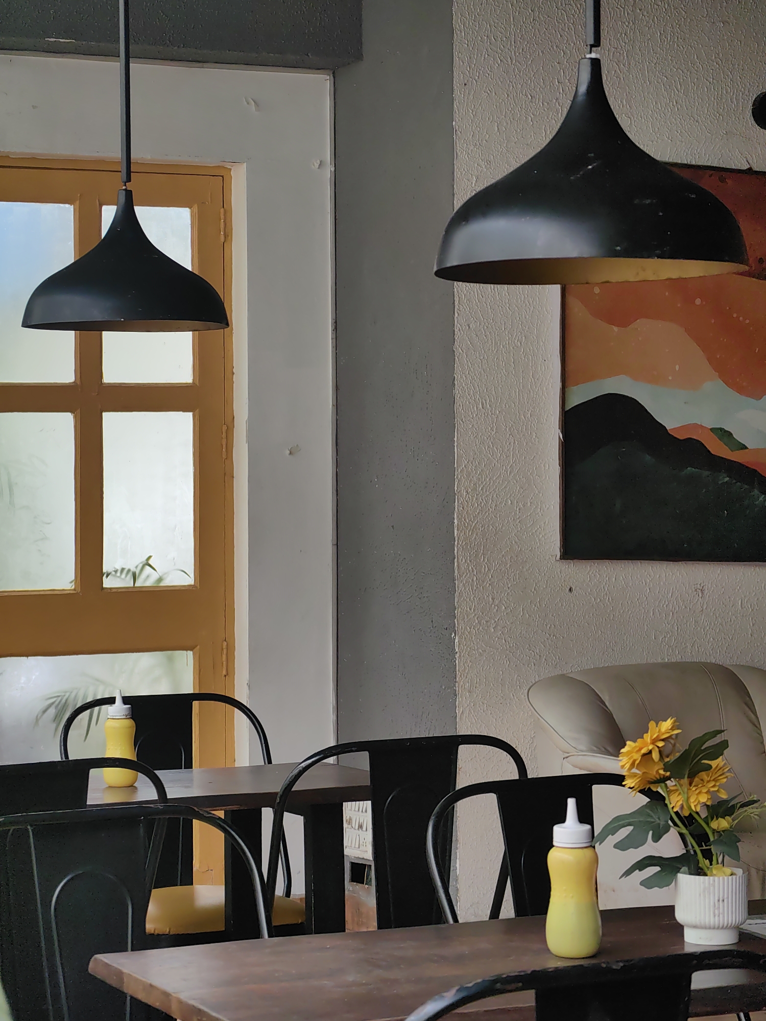 The image captures an interior scene, possibly a cafe, with two black pendant lights hanging from the ceiling. A yellow-framed door with frosted glass panes is visible in the background. Several dark metal chairs with yellow seat cushions surround wooden tables. On one table, a yellow squeeze bottle, likely containing a condiment, and a white pot with yellow artificial flowers are placed. A large abstract painting with orange, green, and black elements hangs on a textured cream-colored wall. A portion of a light-colored leather sofa is also visible. The overall impression is one of a contemporary, minimalist setting with a touch of warmth from the flowers and the painting.