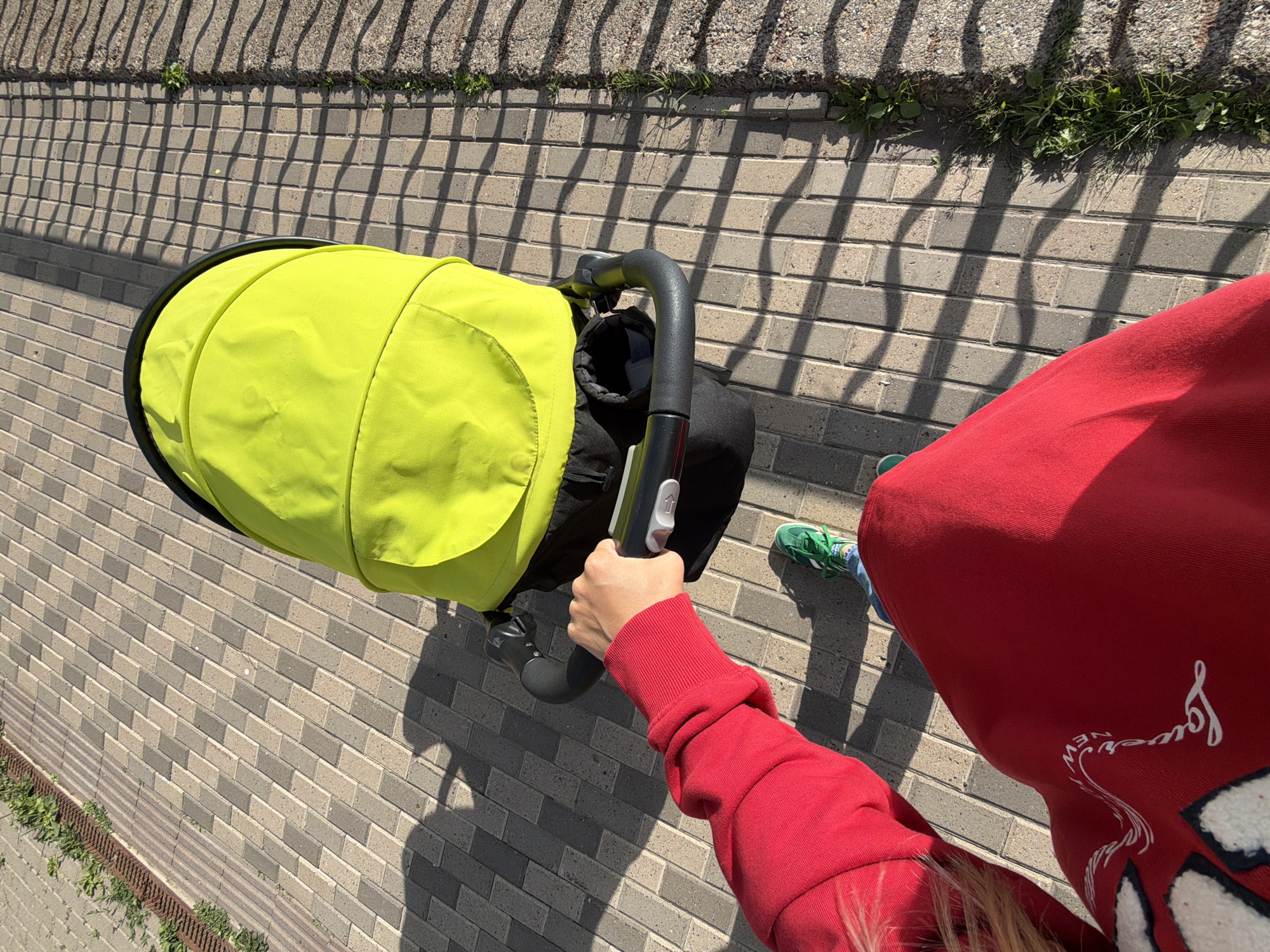 A person's arm in a red sweatshirt is pushing a bright lime green baby stroller on a paved walkway. The stroller has a black frame and a black fabric seat. The person is wearing blue jeans and green sneakers. The walkway is made of rectangular gray and beige bricks. To the right of the walkway is a gravel area with some grass and weeds, bordered by a metal fence casting a shadow. The sun is casting strong shadows on the ground. The scene appears to be outdoors in a city.