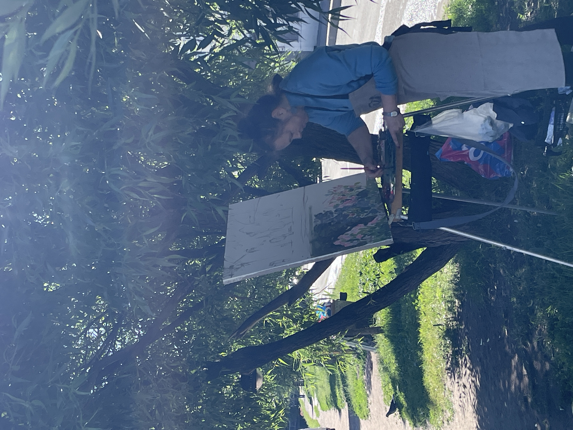 A person is painting outdoors, with a canvas set up on an easel. The painter is wearing a blue long-sleeved shirt and a light-colored apron. They have dark hair pulled back and are focused on the artwork. The painting on the easel appears to be a landscape with flowers.
The scene is set amongst lush green trees, suggesting a park or garden. Sunlight filters through the leaves, creating dappled shadows on the ground. A pathway or road is visible in the background, with grass and trees lining it. There are some bags and supplies next to the easel. The overall atmosphere seems peaceful and creative, indicative of a pleasant day for outdoor painting. The lighting suggests it is daytime, likely mid-morning or afternoon, with clear weather. The location context of Saint Petersburg, Russia, places this scene in a city known for its art and beautiful outdoor spaces.