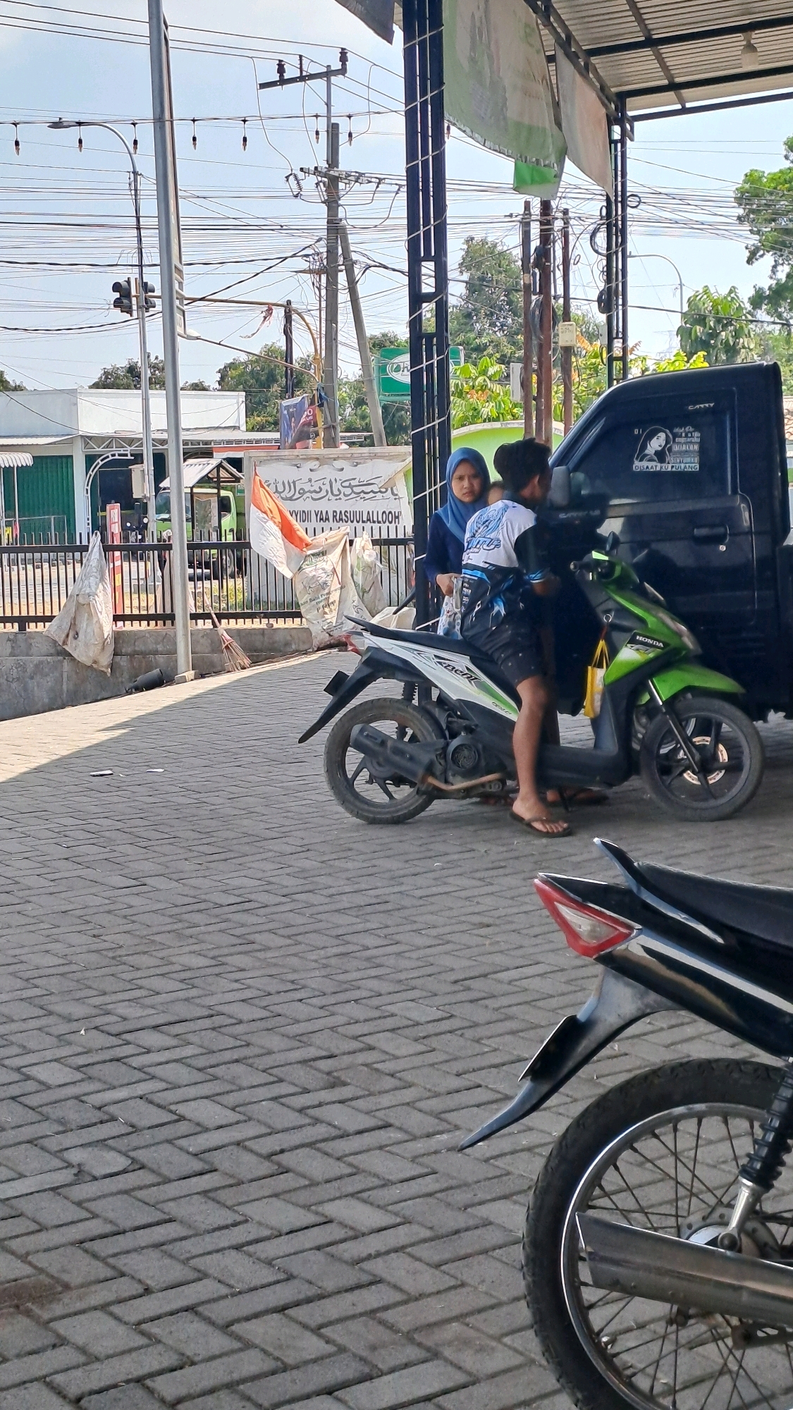 The image depicts an outdoor scene in Kenduruan, Indonesia. In the foreground, a paved area with interlocking brick pavers is visible, with the rear wheel and part of the frame of a dark-colored motorcycle in the lower right corner. In the middle ground, a young person wearing a blue hijab and a young person in shorts and a patterned shirt are standing near a green and white motorcycle. The young person in the patterned shirt is seated on the motorcycle, which is positioned in front of a black pickup truck. Behind the motorcycle and truck, a fence runs horizontally across the image. Beyond the fence, a white banner with Arabic text and an Indonesian flag are displayed. Electrical wires and poles are prominent in the background, suggesting an urban or semi-urban setting. Trees and buildings are also visible in the distance. The lighting suggests it is daytime, with the sun casting shadows.