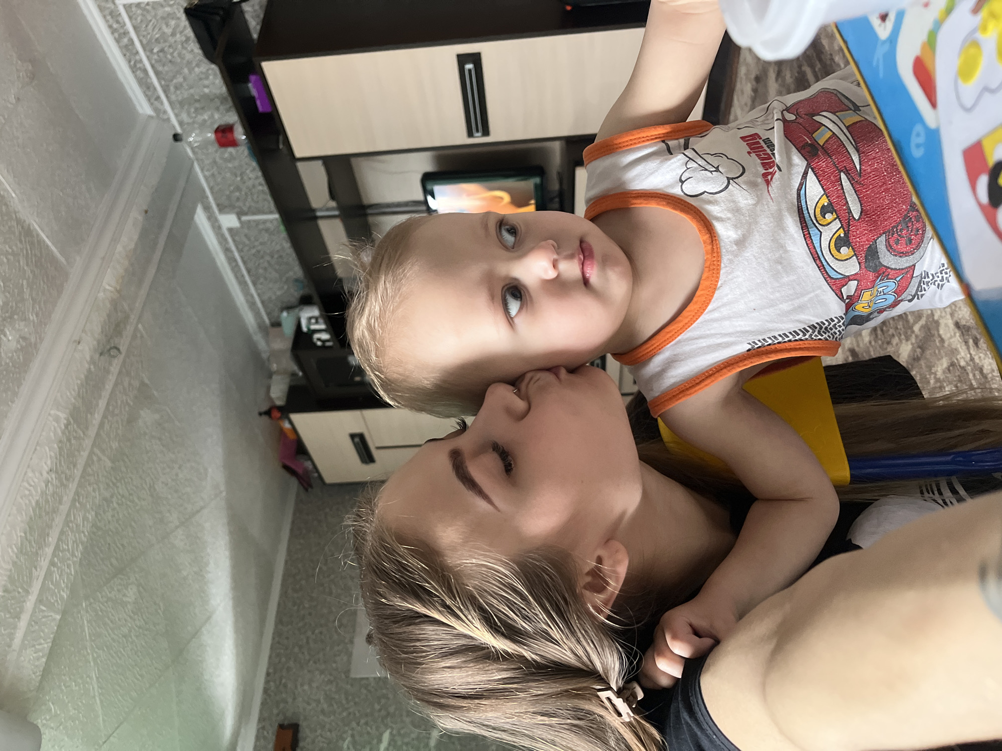 A woman is kissing a young child on the cheek. The child is wearing a white tank top with a red car design and an orange trim. The woman has long brown hair. They appear to be inside a house. A cabinet is behind them. The location is Фоки, Russia.