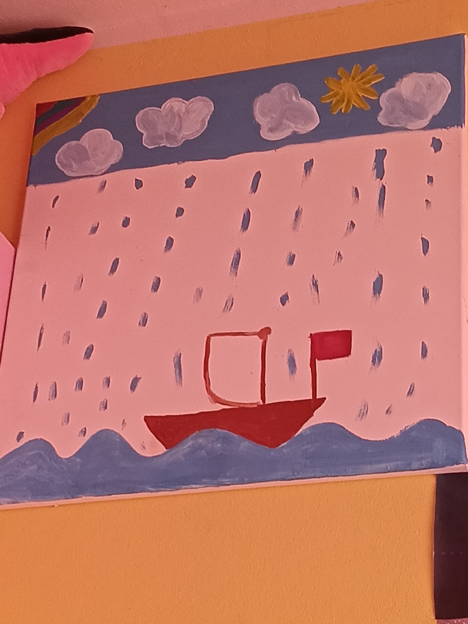 The image displays a painting on what appears to be a canvas, featuring a stylized depiction of a seascape. The painting is positioned on a yellow wall, with a portion of a pink object visible in the top left corner, possibly part of a stuffed animal. The painting itself is divided into sections: the sky is a light blue with white clouds and a yellow starburst. Below the sky is a light pink area with vertical blue strokes, representing rain. In the lower portion of the painting, there are blue wavy lines indicating water, and a red boat with a red flag is sailing on the water. The style is simplistic and childlike. The location is Wels, Austria.