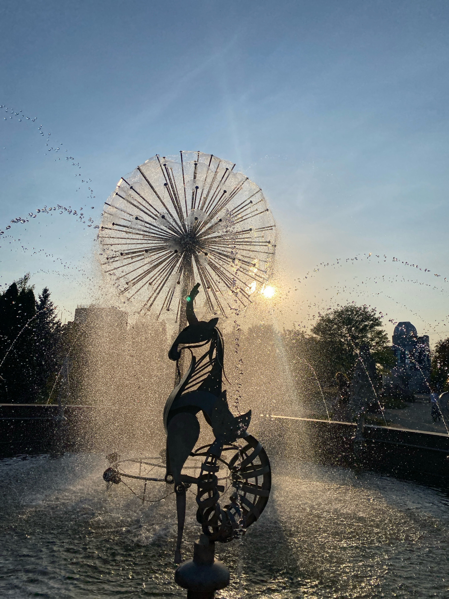 This is a photograph of a fountain with a sculpture in the foreground, set against a backdrop of trees and a building under a clear sky at sunset. The fountain is a complex structure with a large, spherical metal sculpture resembling a dandelion seed head, with thin rods radiating outwards. Water sprays from this sculpture, creating a shimmering curtain. Below the spherical sculpture is a dark, silhouetted metal artwork depicting a figure, possibly an animal, balanced on a bicycle-like mechanism with a spinning wheel. The sculpture is positioned in the center of the fountain's basin, which is filled with water. The water is actively spraying and creating a mist that catches the golden light of the setting sun, which is visible peeking through the spray and behind some trees. In the background, there are trees and a building, with hints of other fountain elements or sculptures further back. The overall scene evokes a sense of motion and light play. This fountain is located in Parcul Catedralei in Chișinău, Moldova.