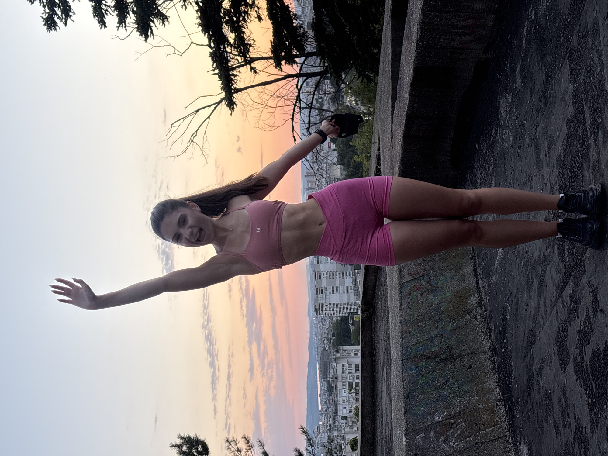 An athletic woman with a toned physique stands on a concrete platform overlooking the city of Varna, Bulgaria, at dusk. She wears a pink sports bra and matching pink shorts, with black athletic shoes. Her right arm is raised in a waving gesture, and her mouth is open as if speaking or shouting. In her left hand, she holds a black kettlebell. The background features a cityscape with numerous buildings bathed in the soft light of a sunset, with a hint of pink and orange in the sky. Trees frame the right side of the image, and a concrete barrier with graffiti is visible in the foreground. The ground is a dark, wet-looking concrete surface.