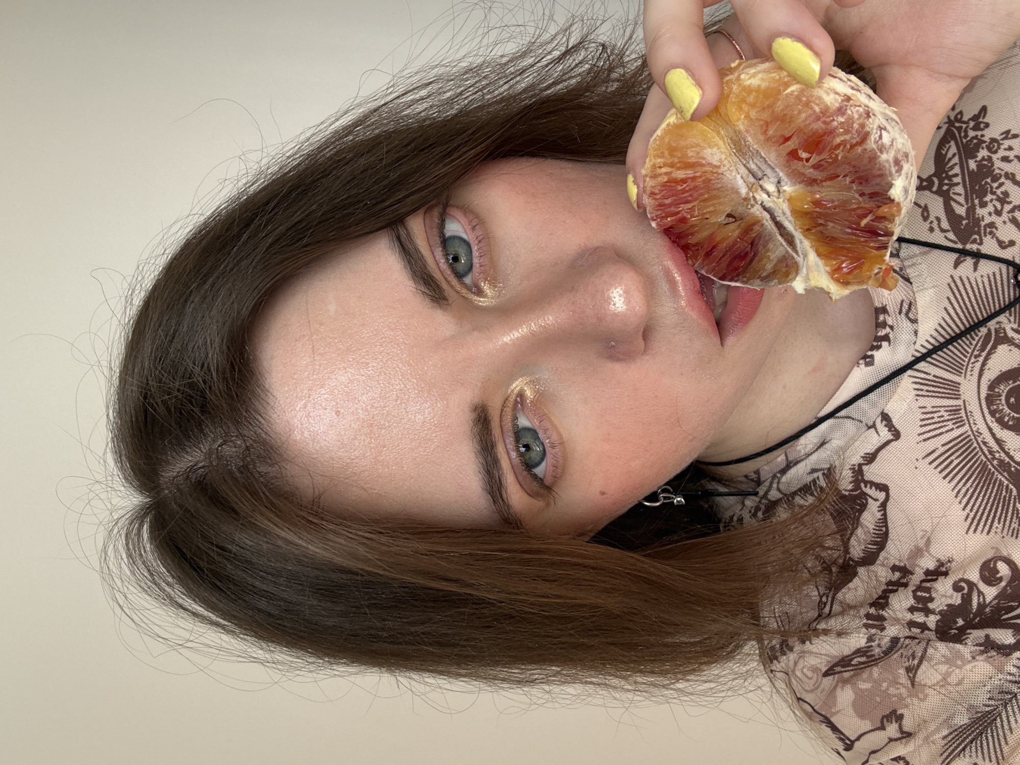 A woman with fair skin and light green eyes, her brown hair parted to the side, is holding a half-peeled blood orange to her lips. Her fingernails are painted yellow. She is wearing a light-colored patterned top. The background is plain off-white. The image does not provide details pertaining to the city of Moscow, Russia.