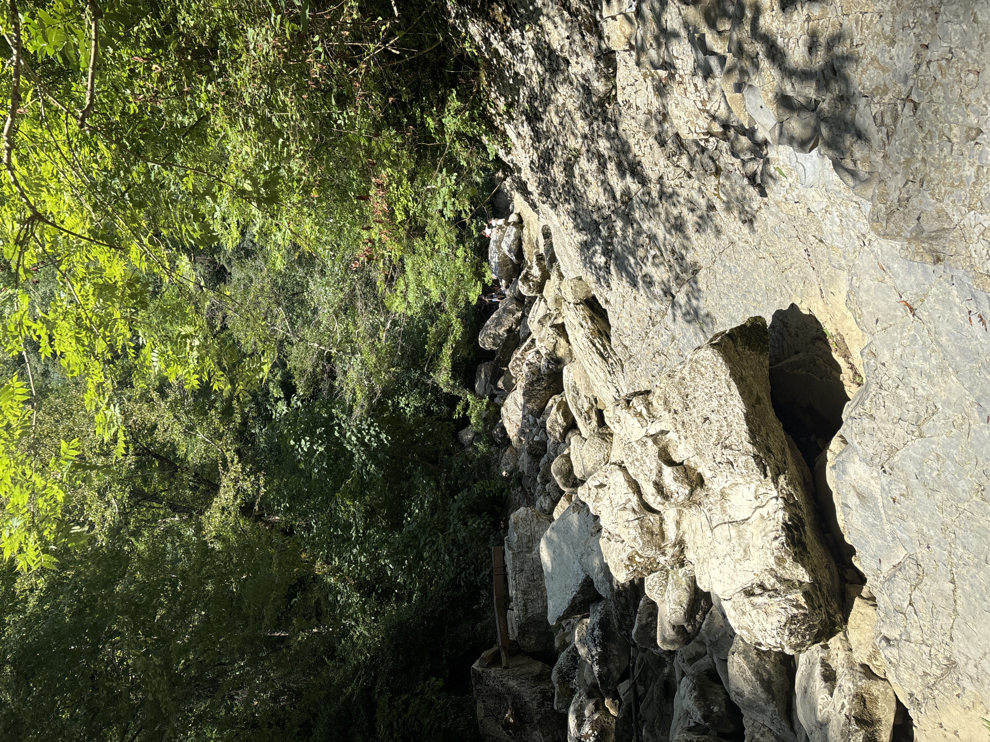 The image shows a rocky, dry riverbed in a wooded area. Large, light-colored boulders are scattered across the landscape, some of them casting shadows from the surrounding trees. The trees are lush and green, indicating a warm season. In the background, a few people are visible walking amongst the rocks. The terrain appears to be rugged and natural. The scene is likely located in a park or natural reserve within Khostinskiy, Russia, given the mention of the city.