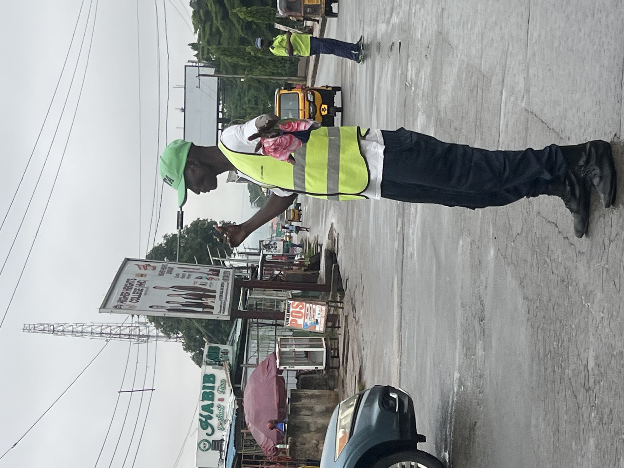 The image captures a street scene in Onitsha, Nigeria, on an overcast day. A Black man wearing a bright green cap and a high-visibility yellow vest over a white t-shirt stands on a wet asphalt road, holding a pink cloth with floral patterns. He is looking to his right, with his left hand raised. His dark pants and black boots are visible. In the background, a yellow auto-rickshaw, also known as a "keke napep," is driving on the road. To the left, a blue car is parked. Further in the background, there are buildings with signs, including one for "HABIB Yoghurt & Fura," and a large billboard for "HIGHER HEIGHTS COLLEGE (HHC)." A telecommunications tower is visible against the sky. Another man in a similar high-visibility vest and dark pants is seen walking in the distance to the right. The overall environment suggests a busy urban setting, likely a market or a commercial area, with signs of recent rain.