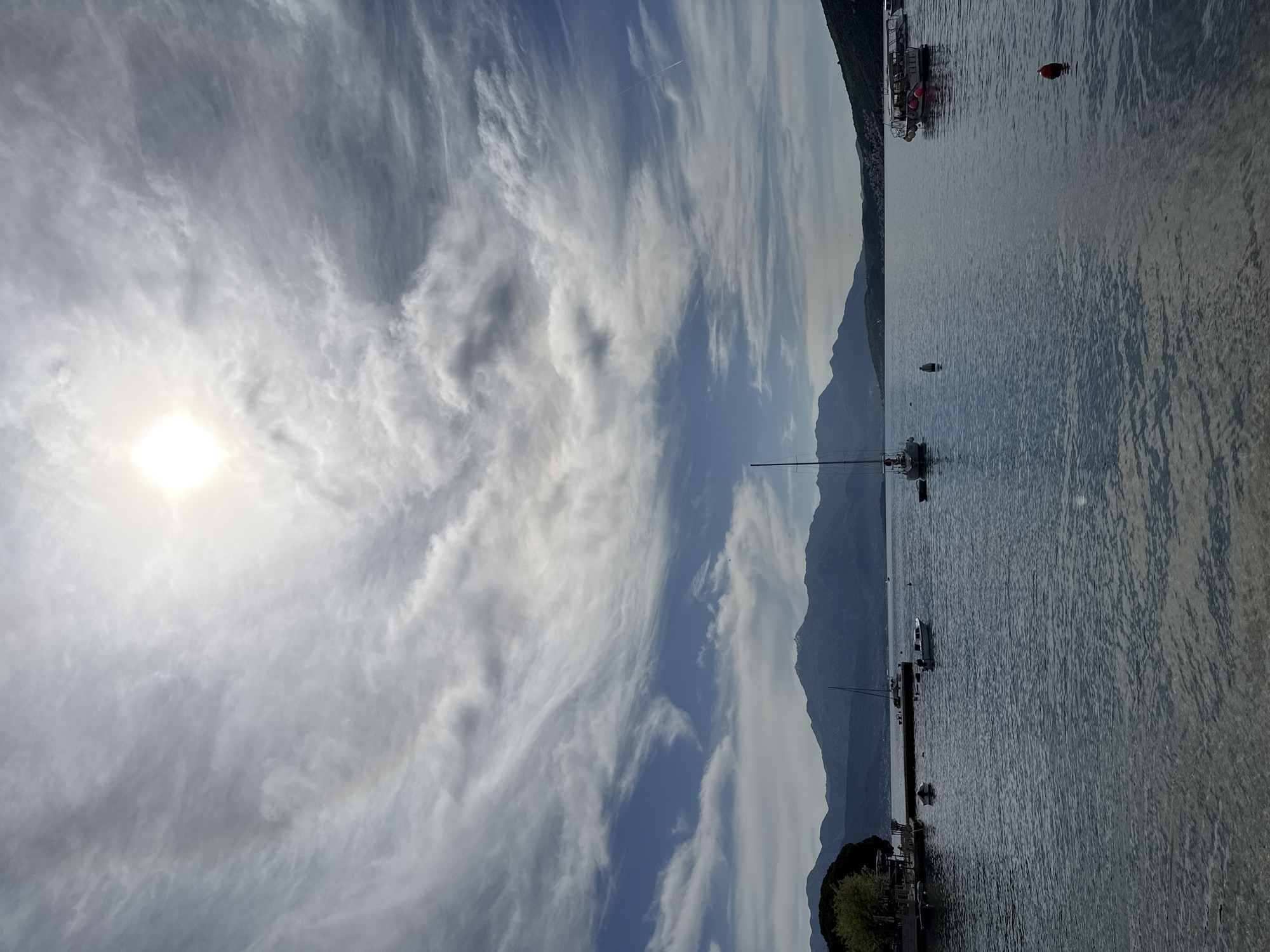 The image displays a tranquil coastal or lakeside scene under a vast, partially cloudy sky, with mountains lining the horizon. The bright sun, partially obscured by wispy clouds, is positioned in the upper left, creating a strong glare and illuminating the expansive skyscape.
The sky is a prominent feature, filled with a diverse array of clouds ranging from delicate, feathery cirrus formations stretching across the upper areas to denser, more textured cumulus clouds below. A faint airplane contrail is visible high in the sky towards the upper right.
The middle and foreground are dominated by a calm body of water, likely a bay or a large lake, exhibiting subtle ripples and reflections. Several boats are scattered across the water: a prominent sailboat with a tall mast is anchored in the mid-ground. Towards the right, a larger boat or ferry with distinct red accents is docked near the shore, accompanied by a floating red buoy. Another smaller, white powerboat is visible further out, and a small boat is anchored closer to the bottom left, near a patch of green foliage indicating land.
In the background, a majestic range of dark mountains rises directly from the water, their peaks appearing somewhat hazy and defining the distant landscape. The scene evokes a sense of peace and natural beauty. The time of day appears to be daytime, possibly morning or late afternoon, given the angle and intensity of the sun. There are no people visible, and no discernible text appears in the image. The setting is consistent with the coastal environment of Đenovići, Montenegro.