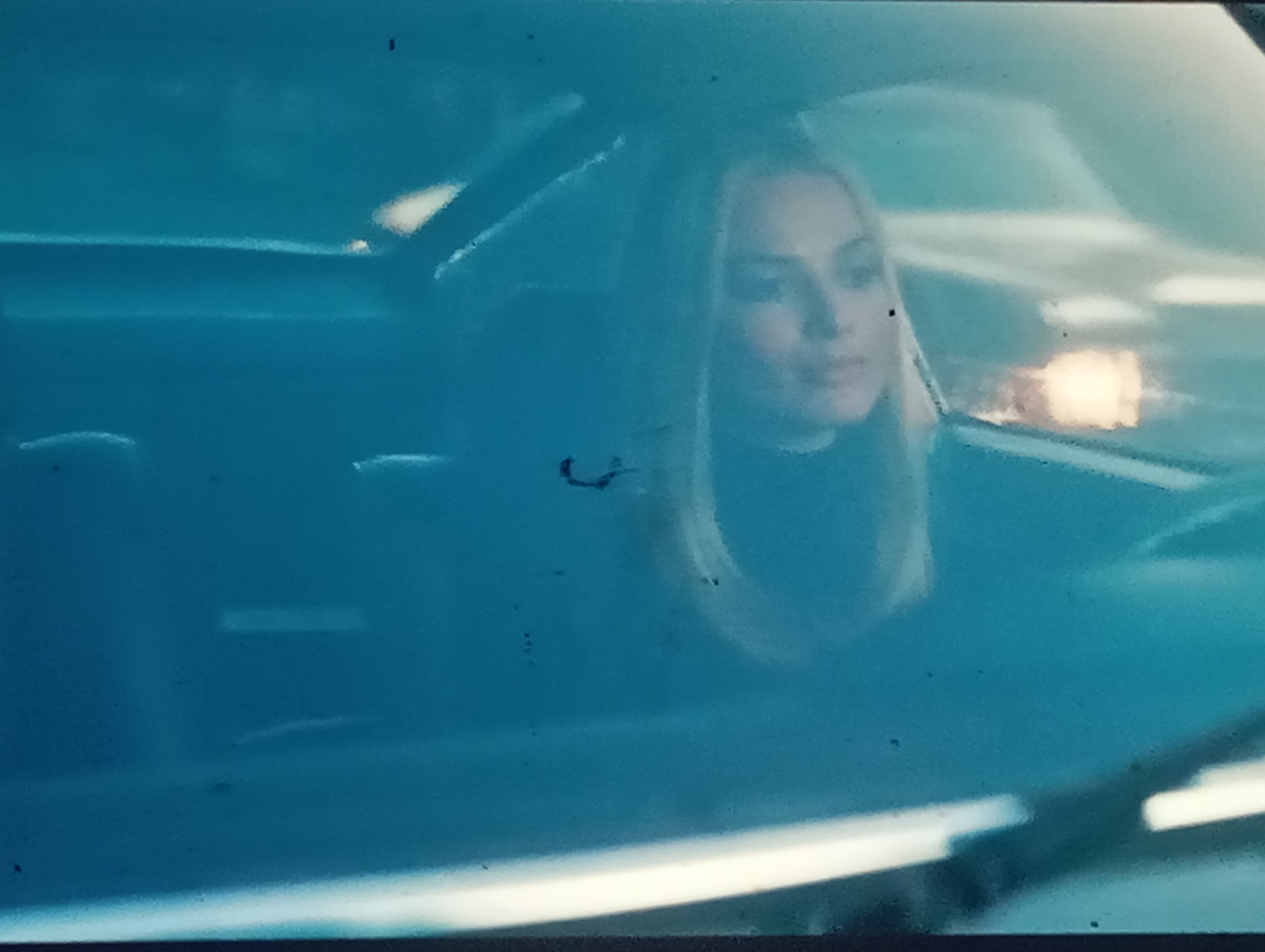 The image shows a woman with blonde hair, wearing a black turtleneck, inside a car. Her face is partially visible through the car window, which is reflecting the surrounding environment. The scene appears to be set at night or in low light conditions, as evidenced by the illuminated lights visible in the reflection. The overall color palette is dominated by blue tones, suggesting it might be a night shot or an artistic choice of lighting. The woman's expression is neutral, and she is looking forward, possibly driving or observing something ahead. The image does not provide any specific clues or context related to Sofiivska Borshchahivka, Ukraine.