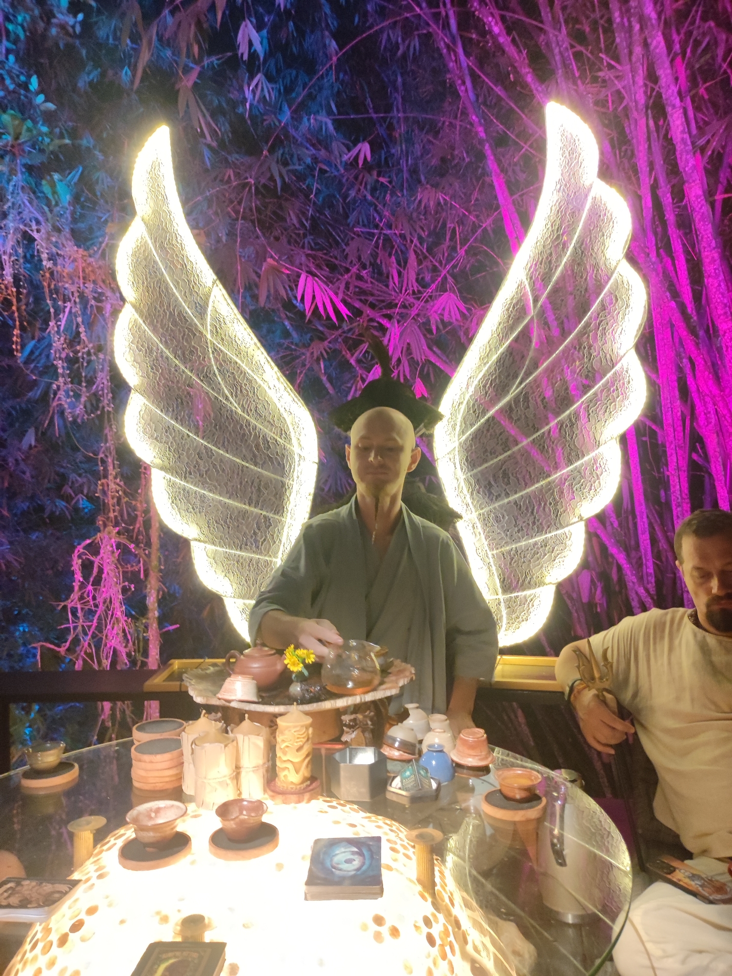The image shows two men seated at a glass table surrounded by various objects. Behind them are illuminated wing-shaped decorations against a backdrop of plants, possibly bamboo, lit with purple and blue hues.
One man, bald and wearing a gray robe-like garment, appears to be preparing or serving tea with a teapot and various cups on a tiered serving tray. The other man is seated to the right, wearing a light-colored shirt.
The table holds numerous items, including stacks of small cups, candles, small containers, and tarot cards. The base of the table seems to be illuminated from within. The scene is set outdoors, likely in a garden or patio area. It suggests a tea ceremony or spiritual gathering of sorts. The location is Lokaserena, Indonesia.