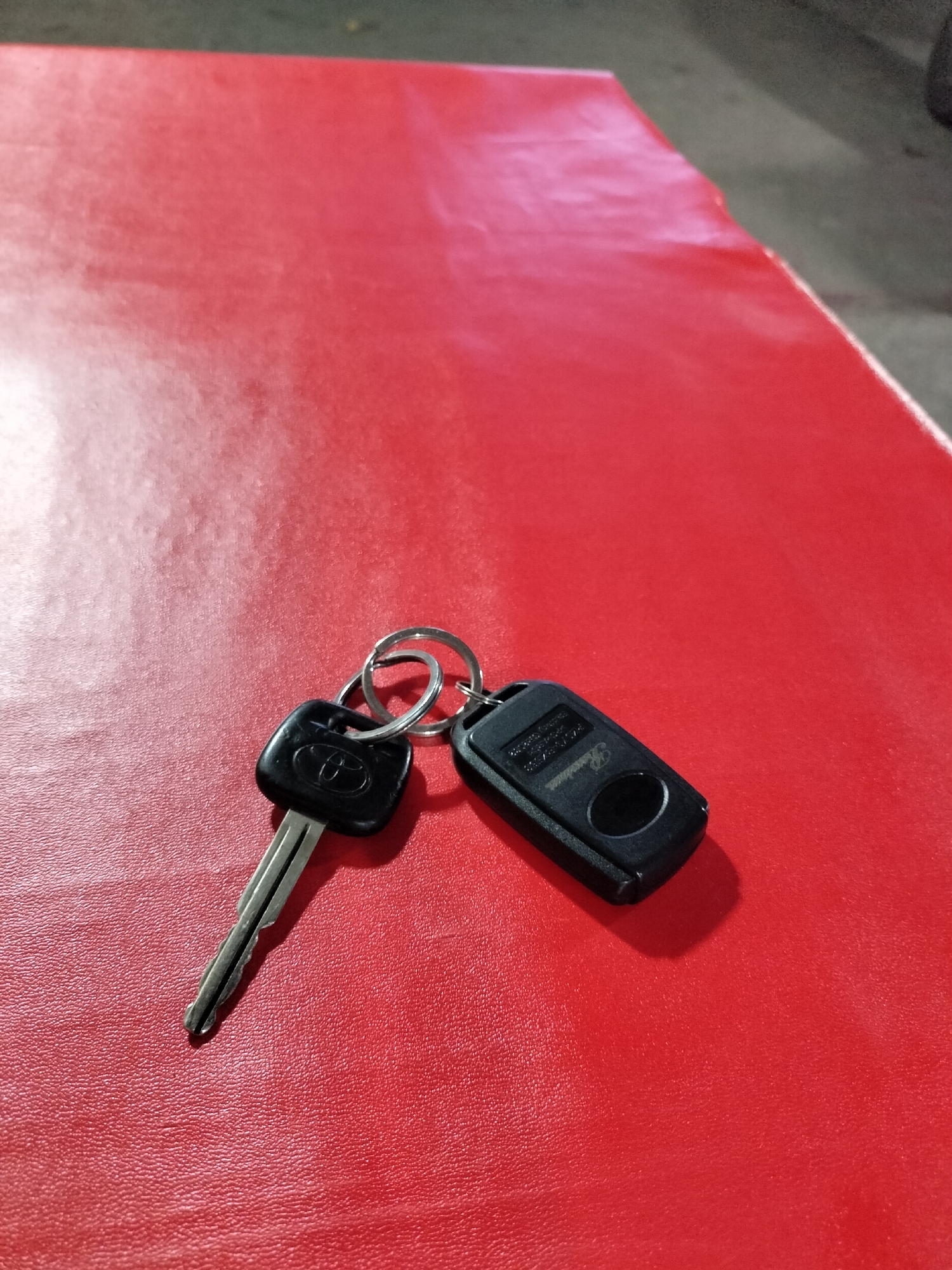 Two car keys, one a standard metal key with a Toyota logo, and the other a black remote fob with a Hyundai logo, are resting on a textured red surface. The red surface appears to be a table or countertop. The keys are attached to a silver keychain ring. The scene is photographed from a slightly elevated angle, with the keys in the lower right quadrant of the frame. The lighting creates subtle shadows on the red surface, indicating a light source from the top left. The background consists of a blurred dark gray surface. The image does not provide any specific visual cues to indicate its location in the city of Langsa, Indonesia.