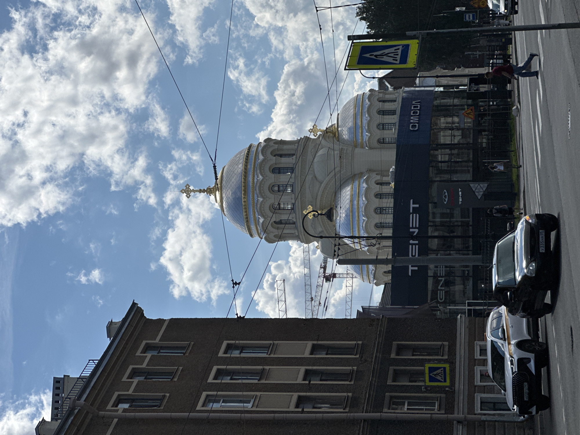 The image captures a street scene in Saint Petersburg, Russia, dominated by the Naval Cathedral of Saint Nicholas. The cathedral, a prominent landmark, features multiple domes topped with crosses, with the largest central dome shining with a golden hue against a partly cloudy blue sky. In the foreground, a multi-story building with a dark facade and numerous windows stands to the left. A busy street with cars and pedestrian crossings runs horizontally across the frame. Several vehicles are visible on the road, including a white SUV and a black sedan. Pedestrians are seen crossing the street and walking on the sidewalk. Construction cranes are also present in the background, suggesting ongoing development or renovation. A large building with the "TENET" and "OMODA" logos is situated in front of the cathedral. Traffic signs, including a pedestrian crossing sign, are strategically placed along the street. Overhead electrical wires crisscross the sky.