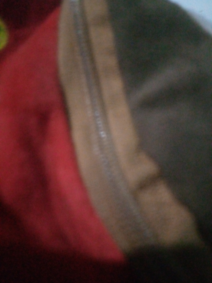 The image is severely out of focus, presenting an abstract, close-up view of textile materials. The left side of the frame is dominated by a blurred red fabric. Adjacent to this, running vertically through the center, is a blurred light brown or tan section, separated from the red by an indistinct, elongated element consistent with a zipper or a prominent seam. The rightmost portion of the image displays a darker, amorphous brown-grey or black material. No individuals, specific objects beyond the textiles, actions, or interactions are discernible. The immediate environment is indistinguishable, and the precise location within the city of Sewon, Indonesia, cannot be determined from the visual content provided.