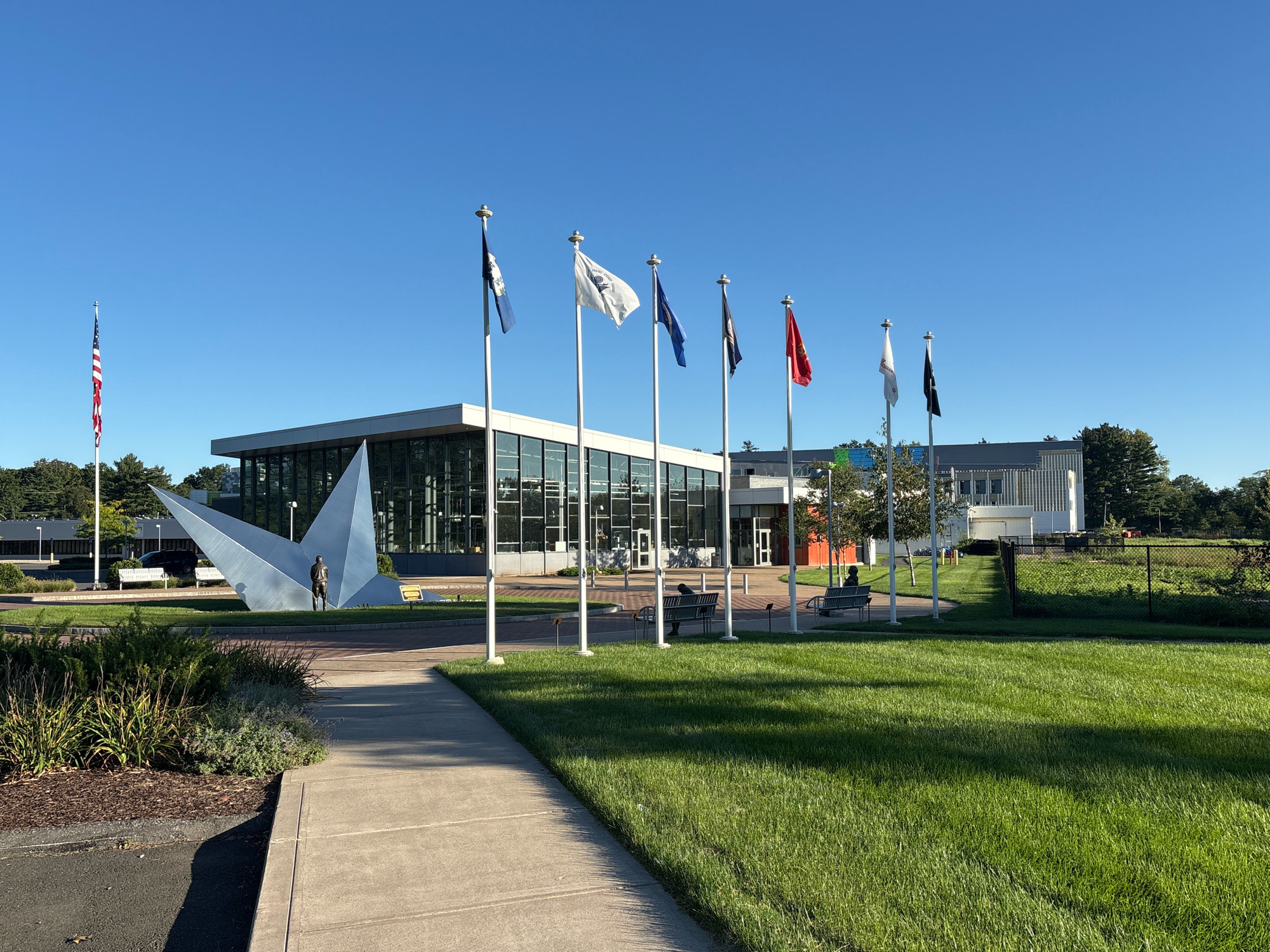 The image depicts the exterior of the U.S. Army Combat Capabilities Development Command (DEVCOM) Soldier Center, located at 1026 St. Nicholas Avenue, East Hartford, Connecticut, United States of America. In the foreground, a concrete pathway runs diagonally across the frame, bordered by manicured green grass on the right and a landscaped garden bed on the left. A large, modern building with extensive glass windows and a white facade dominates the background. In front of the building, several flagpoles stand, displaying various flags, including the American flag on the far left. To the left of the building, a large, abstract metal sculpture with a figure standing in front of it is visible. Several people are seated on benches in front of the building, and a few trees are scattered throughout the scene. The sky is clear and blue, indicating a sunny day.