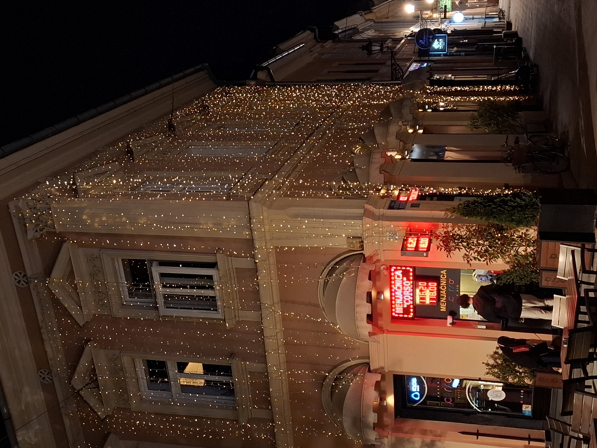 The image captures a street scene at night in Novi Sad, Serbia. An ornate building is decorated with strings of fairy lights stretching across its facade and over the street. Signage visible include "Remier CRAFT BEER HORUS," "MENJAČNICA CORSO," and digital displays with currency exchange rates. People are present in front of the MENJAČNICA CORSO, including one individual who appears to be making a transaction. Tables and chairs are arranged on the sidewalk, and plants are used as decor. A bicycle is leaning against the wall in the middle. The environment is dimly lit, creating a cozy atmosphere.