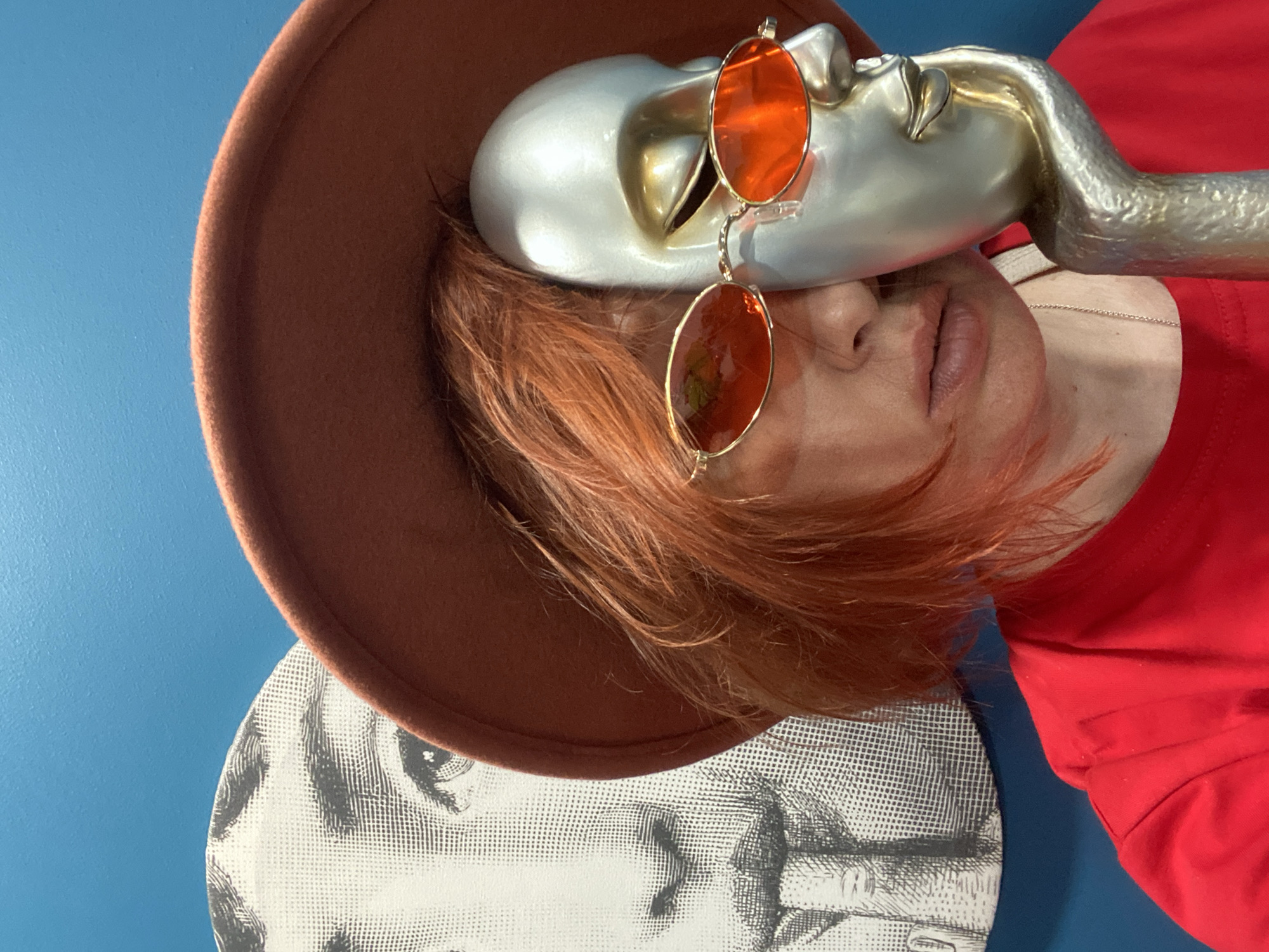 A selfie-style image is taken indoors, likely in a private residence, showcasing an individual with red hair wearing a brown hat. The individual is also wearing orange-tinted round glasses. A silver, abstract face sculpture with attached neck and shoulder rests against the face of the person, partially covering it. The person wears a red top. A round disc with black and white abstract prints is partially visible beneath the hat. The backdrop is a blue wall. The absence of contextual clues makes it impossible to confirm a location in Yekaterinburg, Russia.
