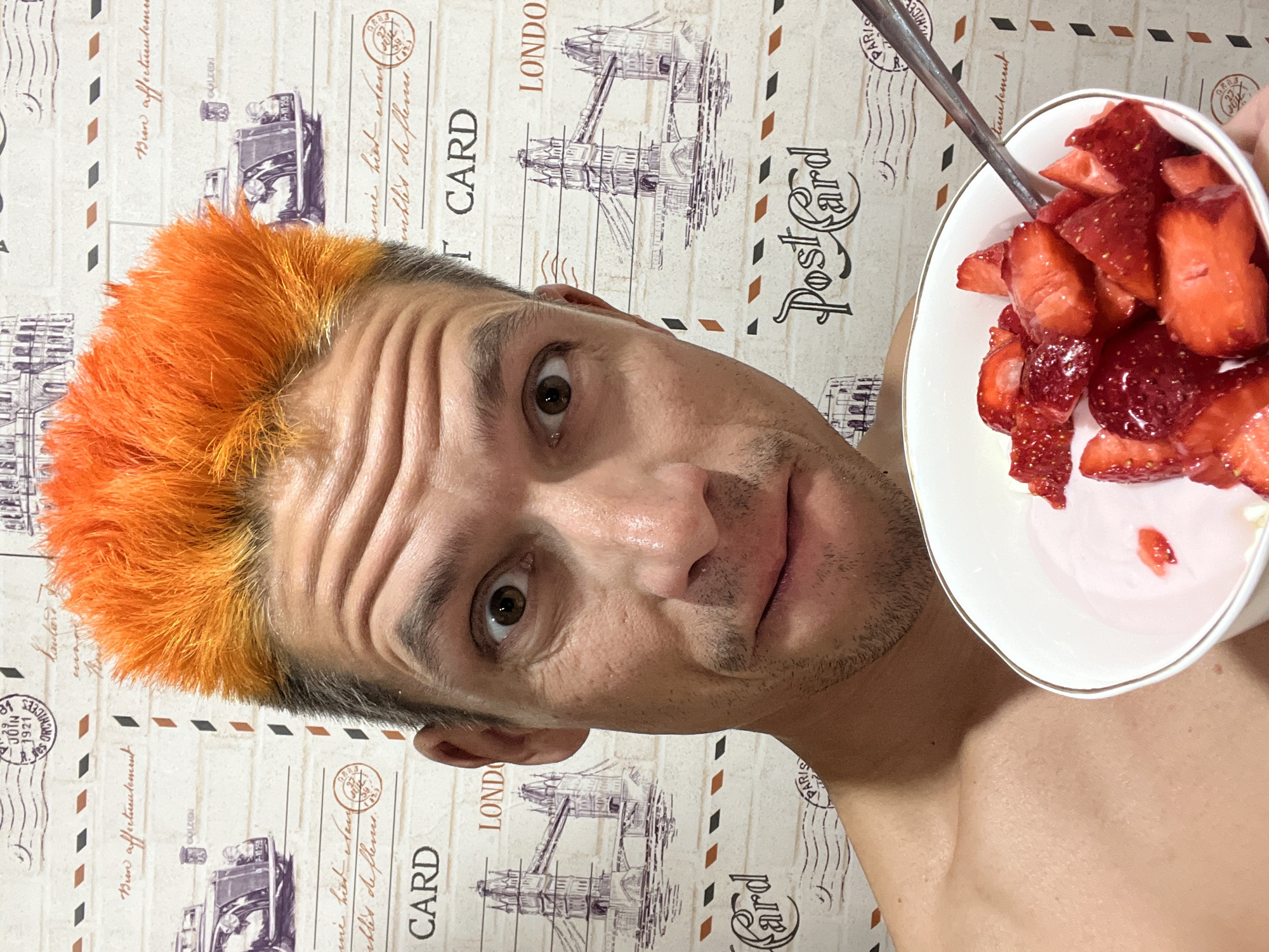 A shirtless man with striking bright orange hair and light stubble is captured in a close-up, angled shot, looking directly at the camera with a wide-eyed, perhaps slightly surprised or engaging expression. His forehead shows several horizontal wrinkles. He holds a white bowl with a gold rim containing chopped fresh strawberries, some mixed with a red liquid, and a silver spoon resting inside. The background is a patterned surface, possibly wallpaper or fabric, designed to resemble vintage postcards. This decorative motif includes illustrations of the Tower Bridge, postmarks (some illegible, others reading "JOIN 1921 ORCHIC"), and text in both English and French. Visible English text includes "CARD," "LONDO" (part of London), and "Post Card." French phrases such as "Bien affectueulement" (Sincerely), "ne hier Hau," and "ibles de fleme" (resembling flowers) are also discernible, along with "PARI" (part of Paris) and "CAALDEN" (likely Golden). Despite the postcard themes hinting at European cities, the actual location context is Chaikovsky, Russia. The indoor setting provides no specific cues for time of day or weather.