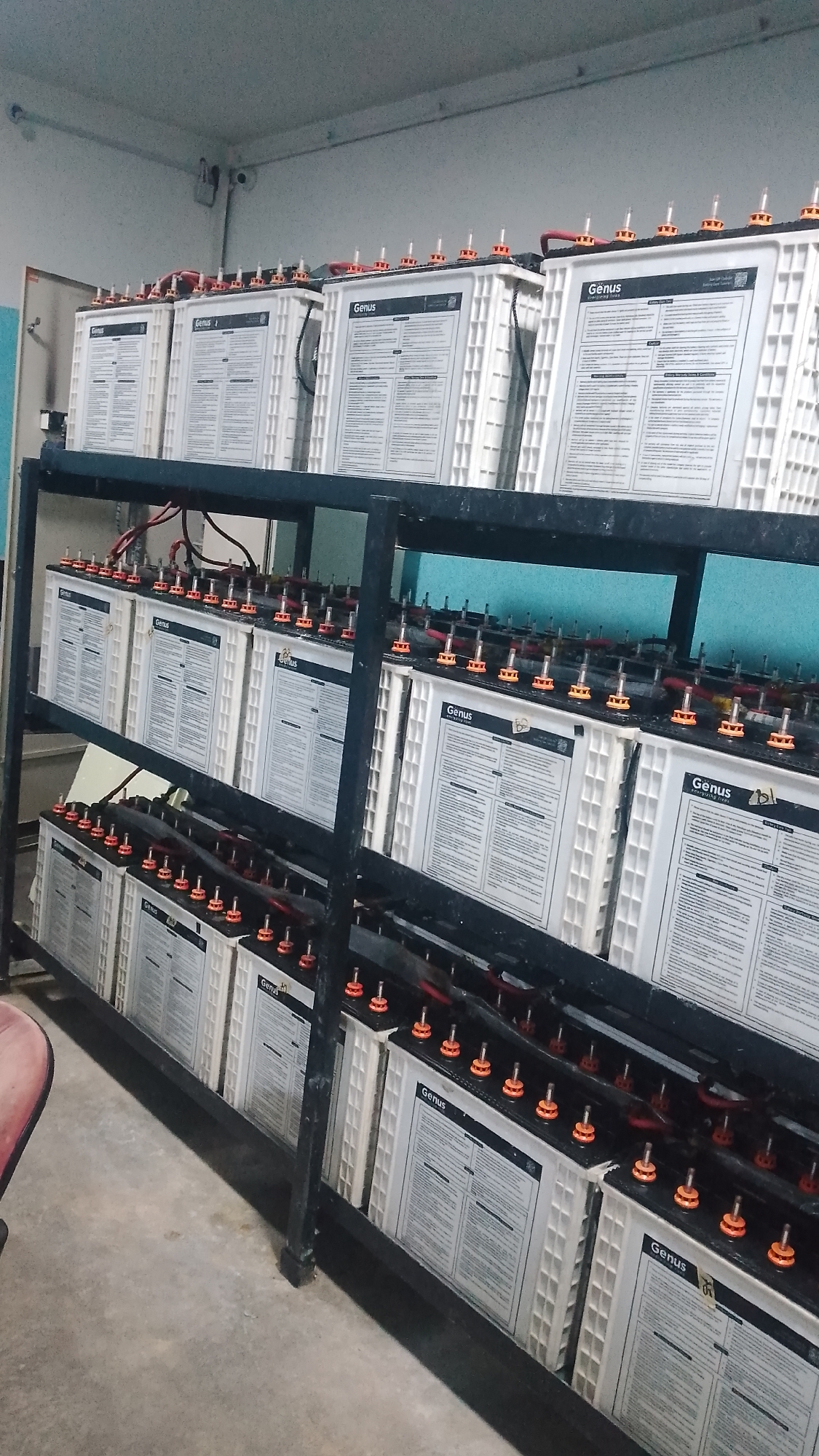 The image depicts a room in Effurun, Nigeria, containing a three-tiered metal rack filled with "Genus" branded batteries. The batteries are white with black wiring connecting their terminals. The room has light-colored walls and a concrete floor. There are electrical conduits visible on the wall.