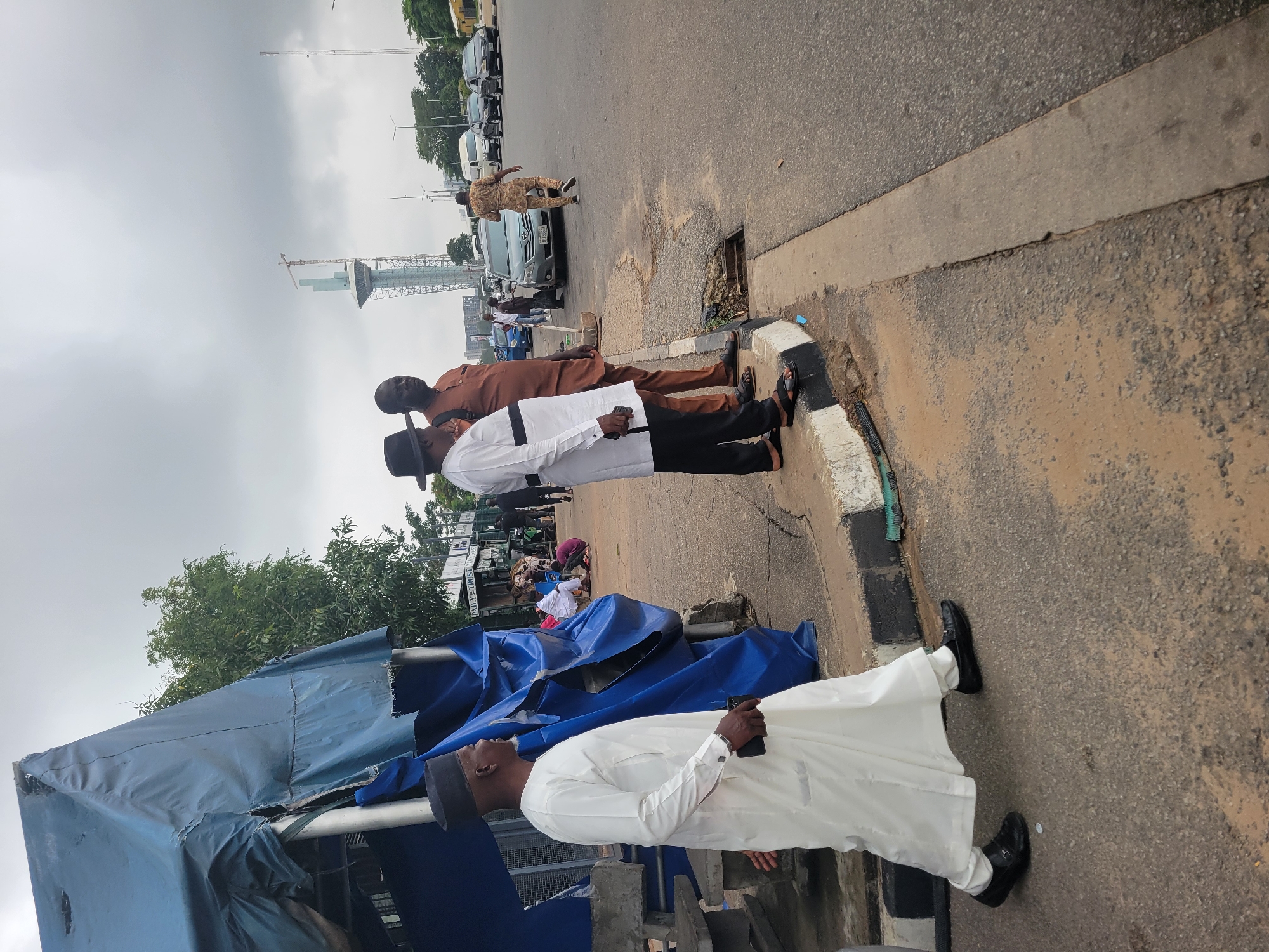 The image shows a street scene in Abuja, Nigeria, under a cloudy sky. In the foreground, a man in a white robe and black cap is walking on a paved sidewalk, holding a black smartphone in his right hand. To his right, two other men are standing on the sidewalk. One is wearing a brown traditional outfit and a black hat, while the other is in a white and black traditional outfit with a black hat. Behind them, a blue tarp is partially covering what appears to be a stall or shelter. In the background, a tall, unfinished skyscraper with a crane is visible, along with other buildings and a busy road with several cars and vans. The sidewalk has a painted curb and some debris. The overall environment suggests an urban setting with ongoing construction and daily life.