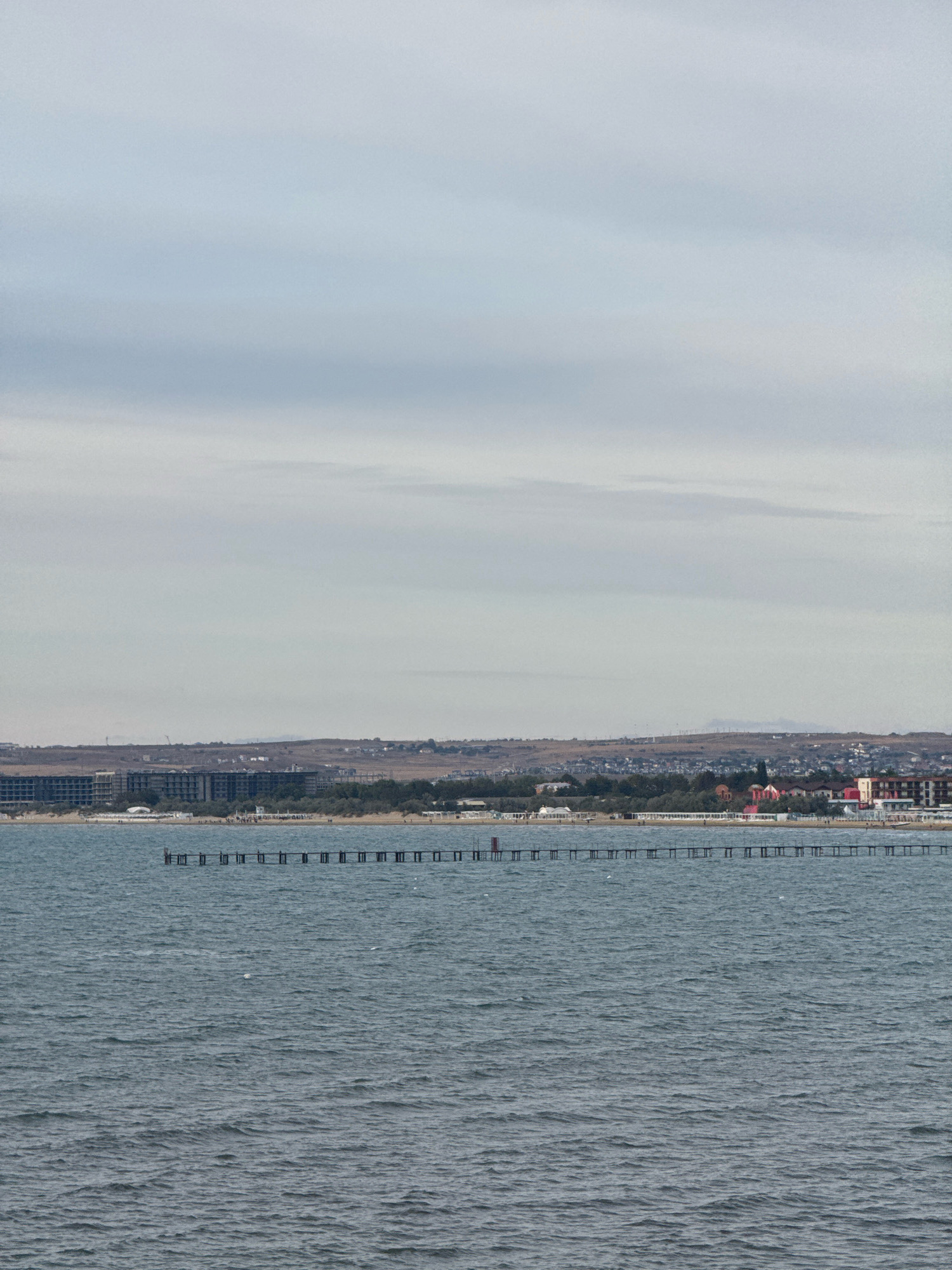 The image depicts a wide view of the Anapa Bay in Anapa, Russia, under a cloudy sky. The foreground is dominated by the dark blue, choppy waters of the bay, with subtle ripples and small whitecaps indicating a moderate breeze. In the mid-ground, a long, wooden pier extends out into the water, supported by numerous pilings. The pier appears weathered, with some sections possibly in disrepair. Beyond the pier, the shoreline of Anapa is visible. A strip of sandy beach is dotted with sparse trees and vegetation. Behind the beach, the city unfolds with a collection of buildings, including several multi-story hotels or apartment complexes with flat roofs and rows of windows. Further inland, the terrain rises into rolling hills, with smaller houses and structures scattered across the landscape. In the distance, the outline of mountains or larger hills is faintly discernible against the hazy horizon. A few white birds are visible in the water. The overall impression is a somewhat bleak, overcast day at a coastal city.