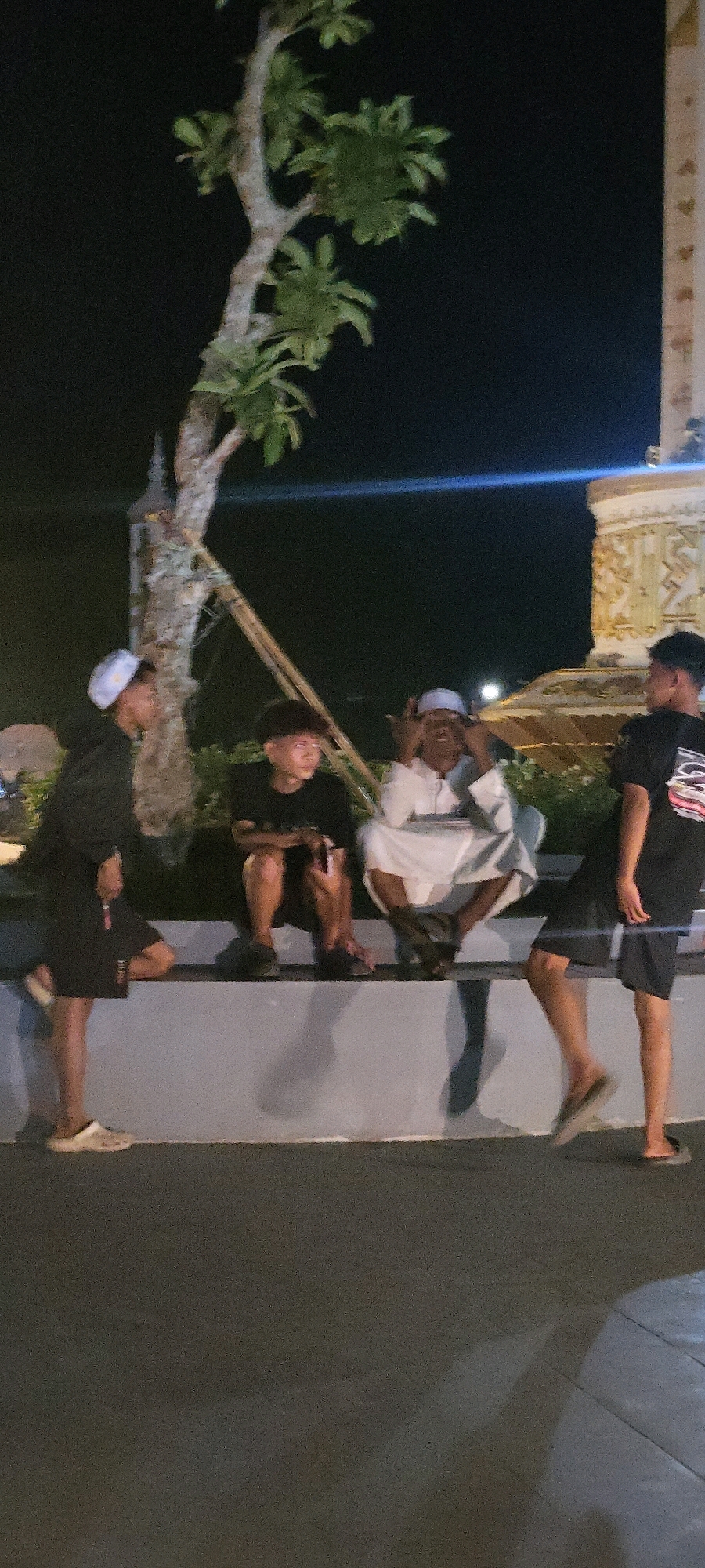 Four individuals are present in the frame, positioned in a dimly lit outdoor setting at night. A tree with sparse foliage stands to the left, with its trunk supported by bamboo poles. In the background, a large, ornate structure with golden accents and decorative carvings is partially visible, suggesting a cultural or historical landmark. The ground is paved with dark material, and a light-colored wall runs horizontally across the mid-ground, serving as a seating area for three of the individuals.
From left to right:
The first person, on the far left, is a male wearing a black hooded jacket and black shorts. He has a white cap on his head and is standing, leaning slightly on the wall. His right foot is adorned with a beige slip-on shoe, and his left foot is in a white slip-on shoe.
The second person, seated on the wall, is a young male wearing a black t-shirt and black shorts. He has short, dark hair and is looking to his right. His feet are bare.
The third person, seated to the right of the second person, is an older male wearing a white kaftan and a white cap. He is gesturing with both hands towards his face. His legs are crossed, and his feet are bare.
The fourth person, on the far right, is a male wearing a black t-shirt with a graphic print and black shorts. He is standing and facing towards the left, with his legs slightly apart. His feet are bare.
The overall scene appears to be a casual gathering outdoors at night, possibly in a public space or near a notable monument, as indicated by the prominent structure in the background. The individuals seem to be conversing or observing their surroundings.