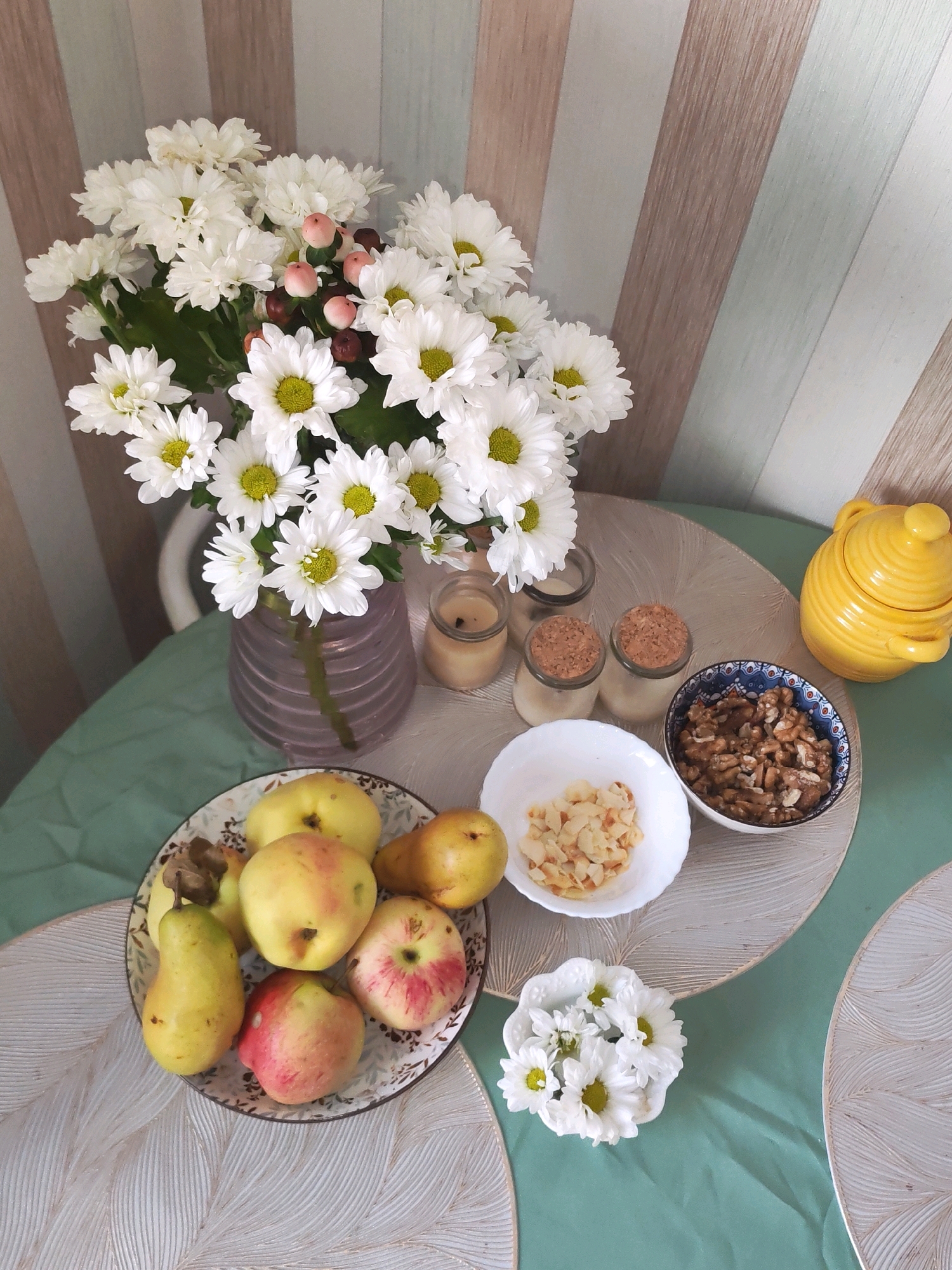 The image depicts a still life arrangement on a table, likely in a domestic setting. In the upper left, a bouquet of white chrysanthemums with green centers is displayed in a purple glass vase. The flowers are interspersed with small, light pink berries and some dark red berries. To the right of the vase, two small glass jars with cork stoppers are visible, containing what appears to be a light-colored substance, possibly honey or jam.
In the center of the table, a white bowl holds slivered almonds. Adjacent to it, a decorative blue and white patterned bowl is filled with walnuts. To the right, a bright yellow ceramic honey pot with a lid and handles is positioned.
In the lower left, a plate with a floral pattern contains a selection of fruits, including yellow apples and pears, as well as red and yellow apples. A single white chrysanthemum in a small decorative dish rests in the lower right of the frame. The table is covered with a light green cloth, and placemats with a textured leaf pattern are visible underneath some of the items. The background consists of a wall with vertical stripes in muted tones of beige, light brown, and light blue-green.
There are no discernible signs indicating a specific location within the city of Izhevsk, Russia. The image is a still life composition, with no people or actions present.