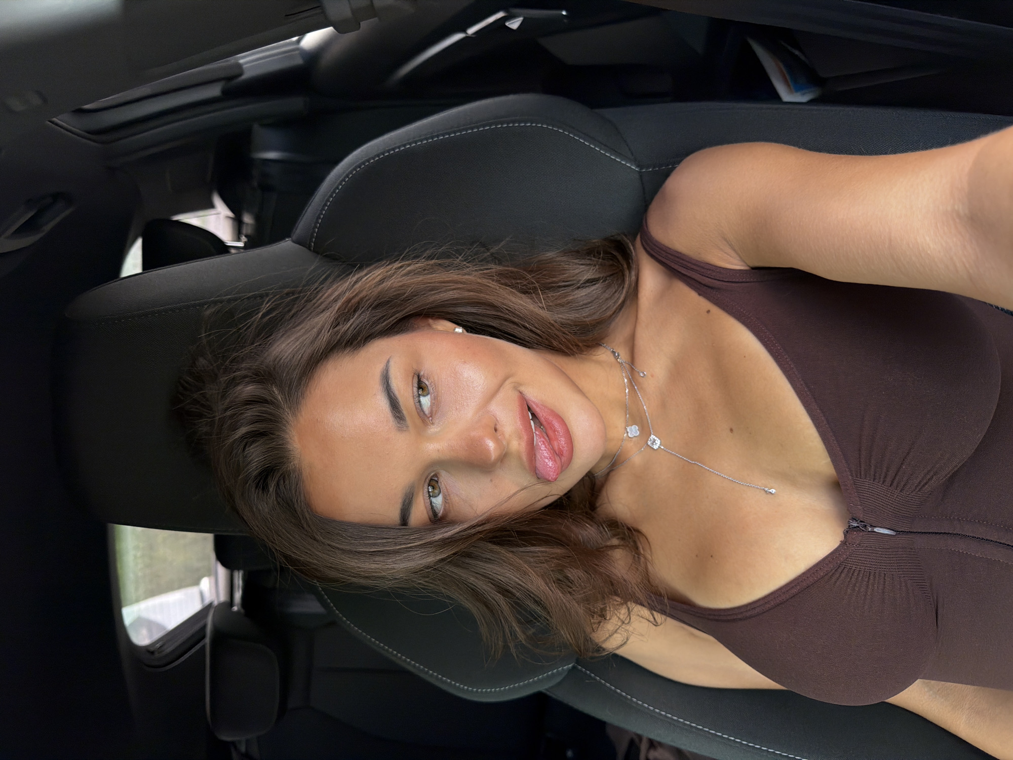 A young woman is lying in the front seat of a car. She is wearing a brown top and a silver necklace. The car has black leather seats and a sunroof. The location of the image is unknown and cannot be determined from the information provided.