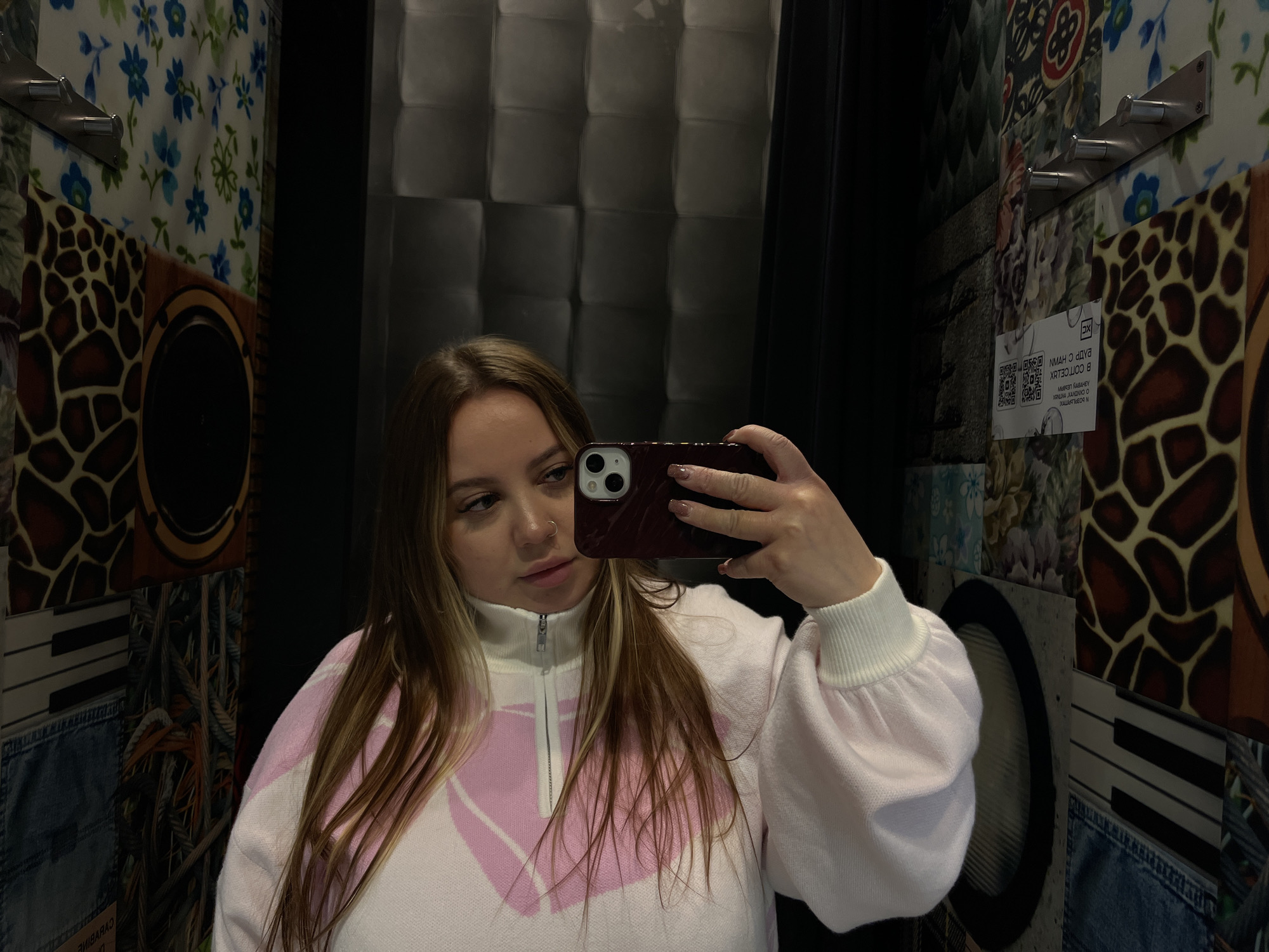 A young woman is depicted taking a mirror selfie indoors. She is positioned slightly to the left of the frame, holding a smartphone with a dark red case in her right hand, which is visible as a reflection. Her light brown hair with blonde highlights falls over her shoulders. She is wearing a white sweater with a pink geometric pattern and a high zipped collar. A nose piercing is visible on her left nostril, and her fingernails are manicured with a light polish.
The background is a strikingly eclectic wall covered in various patterned panels, suggesting a uniquely designed interior, possibly in Krasnodar, Russia. Visible patterns include blue floral motifs on a white background, a brown and black animal print (resembling giraffe or leopard spots), a black and white piano keyboard design, and a denim-like texture. Part of a circular speaker or decorative frame is also seen on the wall. A section of the wall to the upper right features a dark, padded texture.
Several objects are attached to the wall, including a silver wall-mounted rack with three hooks. A white rectangular sign with a QR code and some inverted text is visible on the wall to the right. The visible inverted text includes "В СОРТЕЛХ", "NMAH C YHVN", and "N BОРЧЕFТ". Below the denim pattern, blurry inverted Latin characters are partially visible, possibly spelling "CABARINE". The lighting is artificial, creating some shadows on the woman's face and in the background. The overall impression is an informal moment captured in a distinctively decorated space.