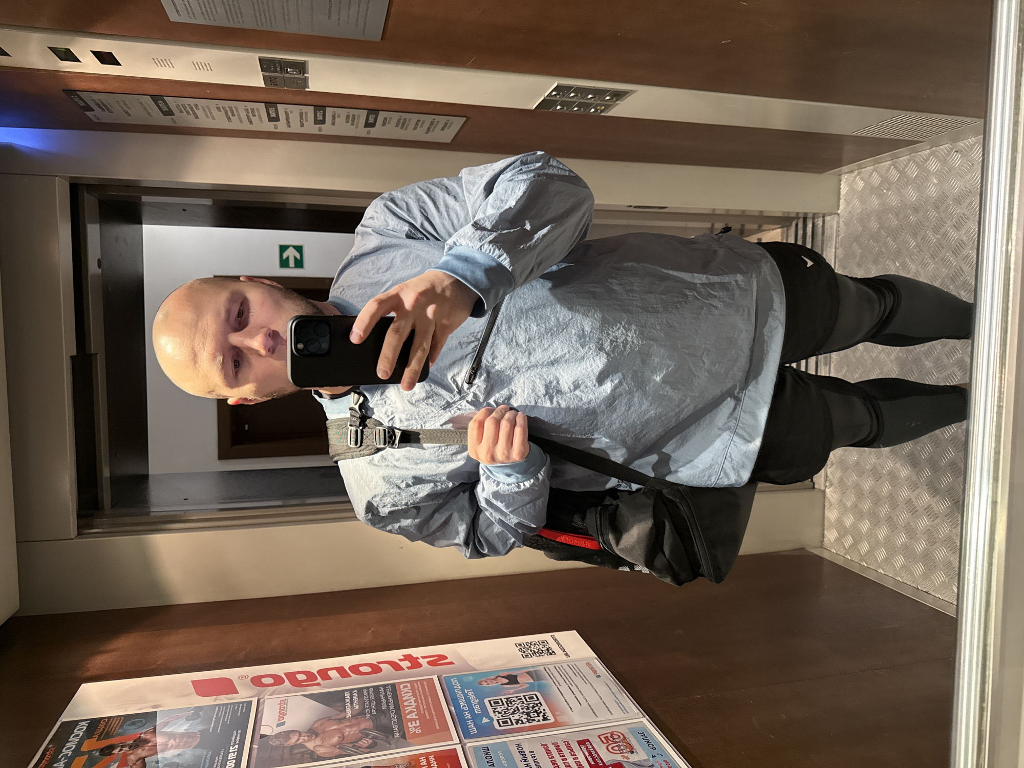 A bald man is taking a mirror selfie in what appears to be an elevator. He is wearing a light blue jacket and black pants, with a black shoulder bag slung across his body. He is holding a black smartphone. The elevator has a metal diamond-patterned floor and the walls are wood-paneled with metal trim. On the floor in front of the mirror are several pamphlets with Russian text, including the word "Strongdo". Based on the pamphlets, the image was likely taken in a Strongdo fitness center, a chain in Krasnoyarsk, Russia.