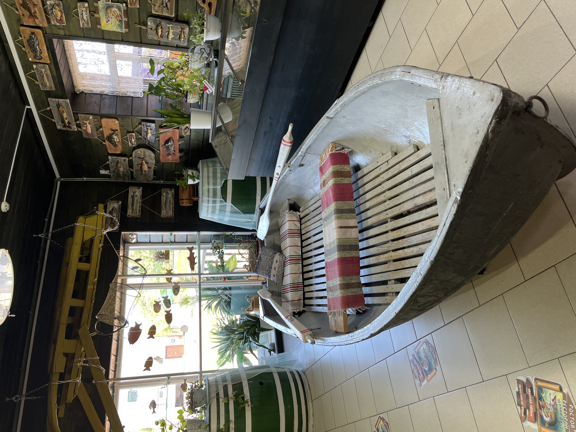 This image depicts the interior of what appears to be a museum or themed establishment, possibly a restaurant, located in Ново-Никольское, Russia. The primary subject is an old, weathered metal rowboat placed centrally on a tiled floor. The boat is equipped with wooden slatted seats and has two decorative striped cushions and throws placed on them. A wooden oar with red and white painted accents rests inside the boat.
The background features walls clad in dark wooden planks, adorned with numerous wooden plaques depicting various types of fish, suggesting a fishing or aquatic theme. A large window with white lace curtains reveals an outdoor scene with greenery, indicating a daytime setting. To the left of the boat, there's a large green barrel decorated with white stripes, and more plants are visible. To the right, a dark display case with glass tops is present, also showcasing plants. The floor is tiled in a light beige color. Several floor decals with images of fish and text in Russian are scattered on the tiles in the foreground, pointing towards the boat. The overall atmosphere is rustic and thematic, emphasizing a connection to fishing and nature. There are no people visible in the image.