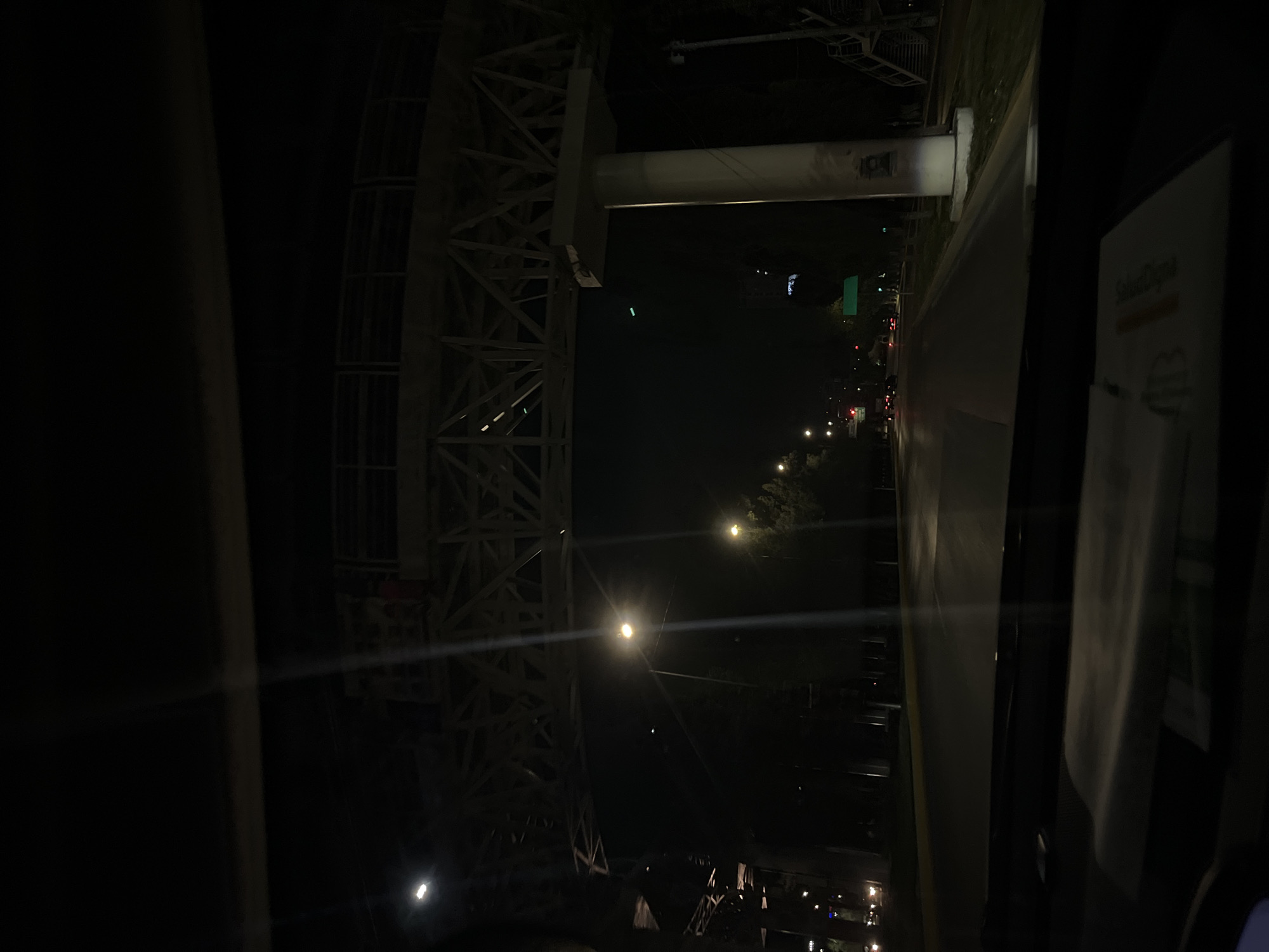 The image depicts an evening view from inside a vehicle in Puebla, Mexico. The scene is dominated by the metal framework of a large bridge, possibly the Distribuidor Vial Zaragoza bridge, illuminated by streetlights. A portion of the bridge's support structure is visible overhead. To the right, a road stretches into the distance, lined with streetlights and possibly other vehicles. The interior of the vehicle is visible on the right edge of the frame, showing part of the dashboard and other interior elements.
