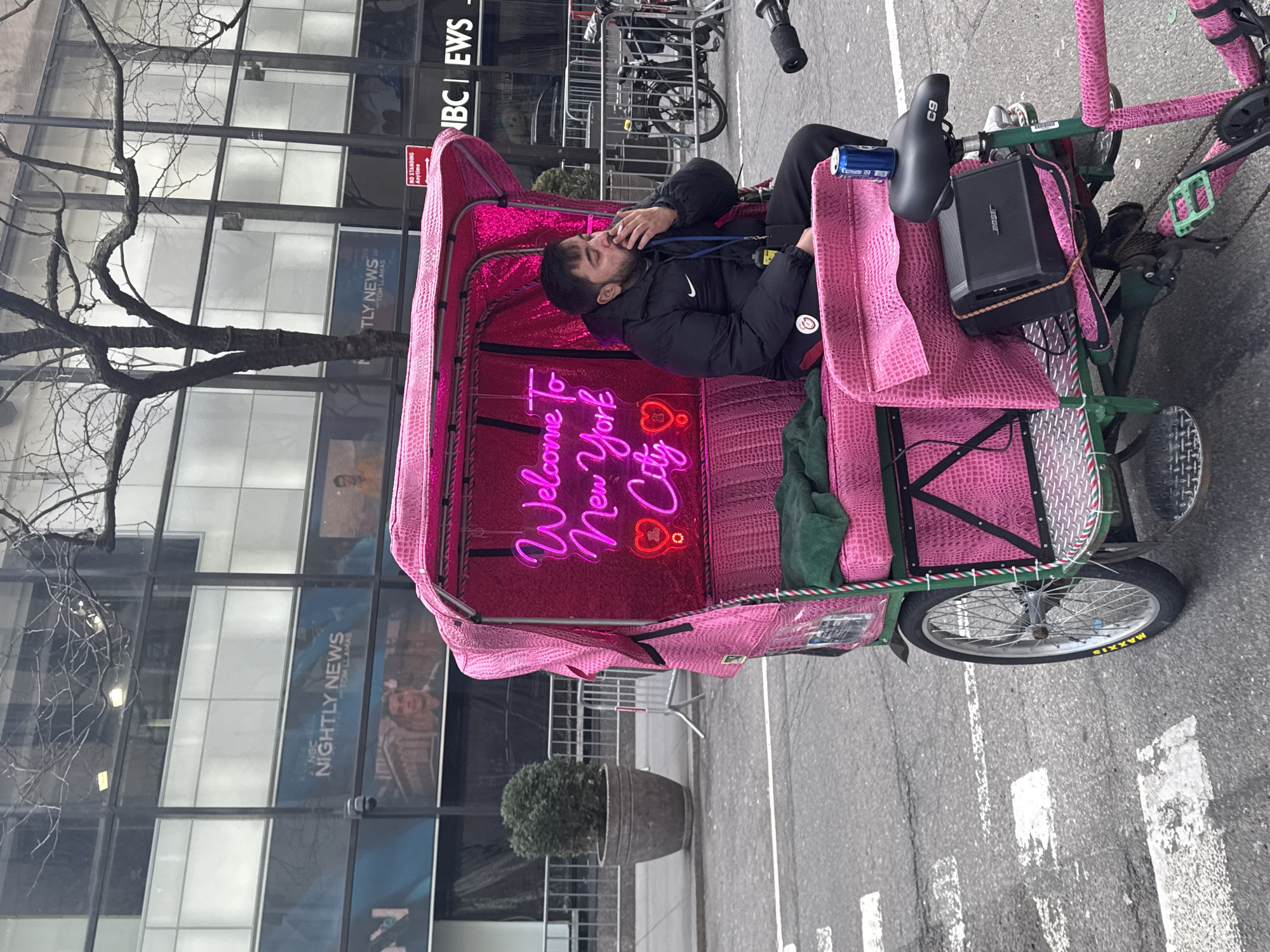 A pink pedicab is parked on an asphalt street in New York City, United States of America, specifically near Rockefeller Center, indicated by "NBC NIGHTLY NEWS" and "NBC NEWS" signage on a background glass building. The pedicab features a pink crocodile-skin textured upholstery, a dark green frame with red and white striped accents, and a glowing pink neon sign inside the passenger area displaying "Welcome To New York City" with two red heart icons. A male individual with dark hair and a beard, wearing a black puffer jacket with a Nike logo and dark pants, is seated in the pedicab's passenger compartment, holding a lit cigarette to his mouth. A blue lanyard with a yellow badge is visible around his neck. A blue beverage can rests on the pedicab's black bicycle seat, and a black Bose portable speaker is secured to its rear frame. The street features white crosswalk markings. The background includes a bare-branched tree, metal crowd control barriers, a potted green plant, and bicycle racks with parked bicycles. A red "NO STANDING Anytime" sign is also present. The scene is under ambient daylight conditions.