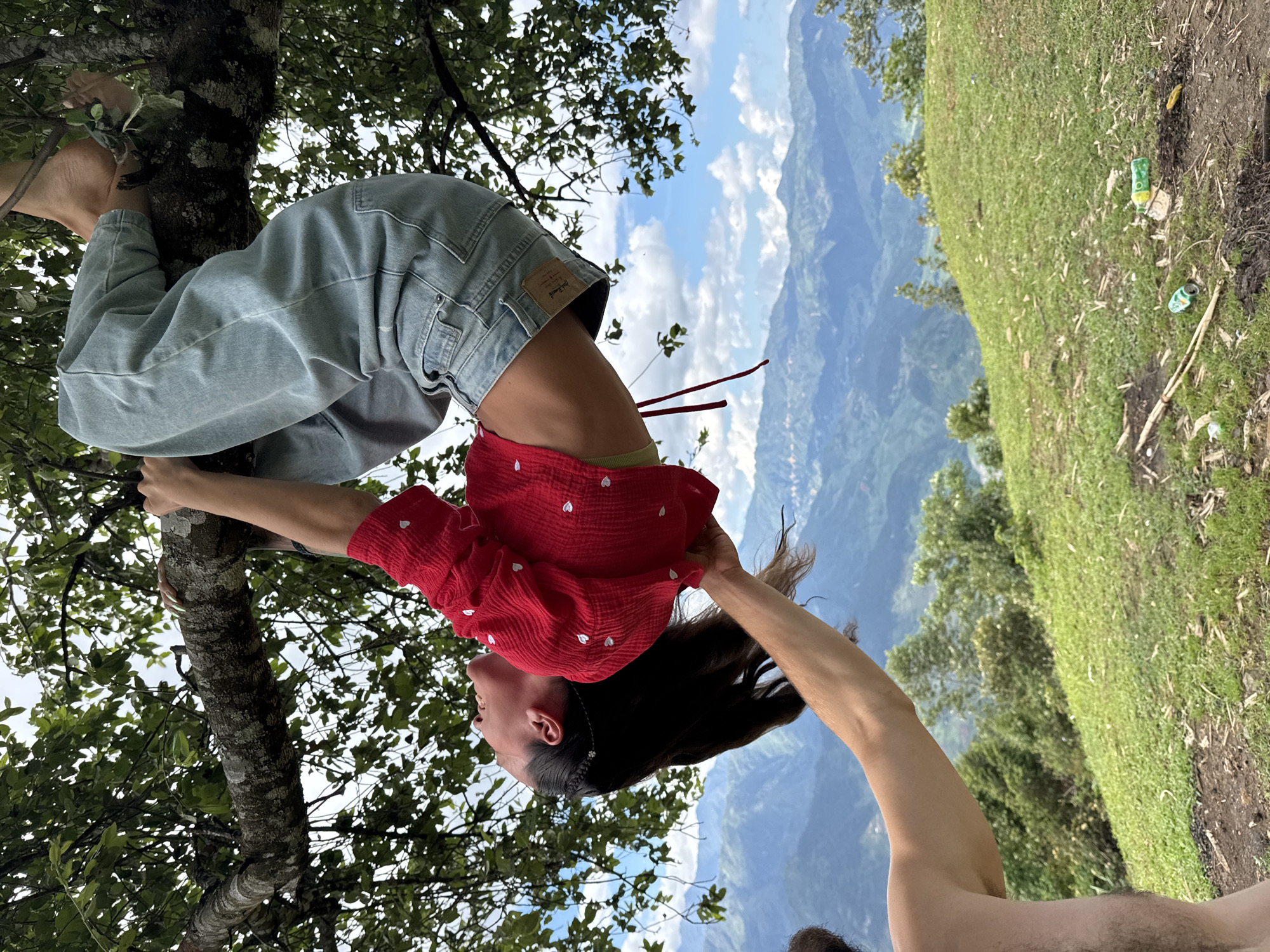 The image displays an outdoor scene in Tú Lệ, Vietnam, featuring two individuals interacting with a tree branch against a mountainous landscape. A female individual is suspended upside down from a thick, horizontally extending tree branch, which is covered in dark bark and green foliage. She wears a red long-sleeved top adorned with small white heart patterns, light blue loose-fitting denim jeans with a brown brand patch on the right rear pocket, and a light green undergarment visible beneath her red top. Her dark hair hangs downwards, and her feet are bare. Her hands grasp the tree branch, and her legs are wrapped over it. Her head is tilted back, looking upwards towards the partly cloudy sky.
A second individual, partially visible in the lower-left quadrant of the frame, extends a bare, hairy arm upwards, holding the left hand/forearm of the suspended female, providing support. The background showcases a series of green, forested mountains receding into the distance, under a sky with visible blue patches and white cumulus clouds. The foreground consists of a green grassy slope with areas of exposed brown earth. Scattered on this ground are several pieces of debris, including at least two light-green beverage cans, multiple light-colored wooden or bamboo fragments, and a yellow object resembling a banana peel or corn cob in the lower right.