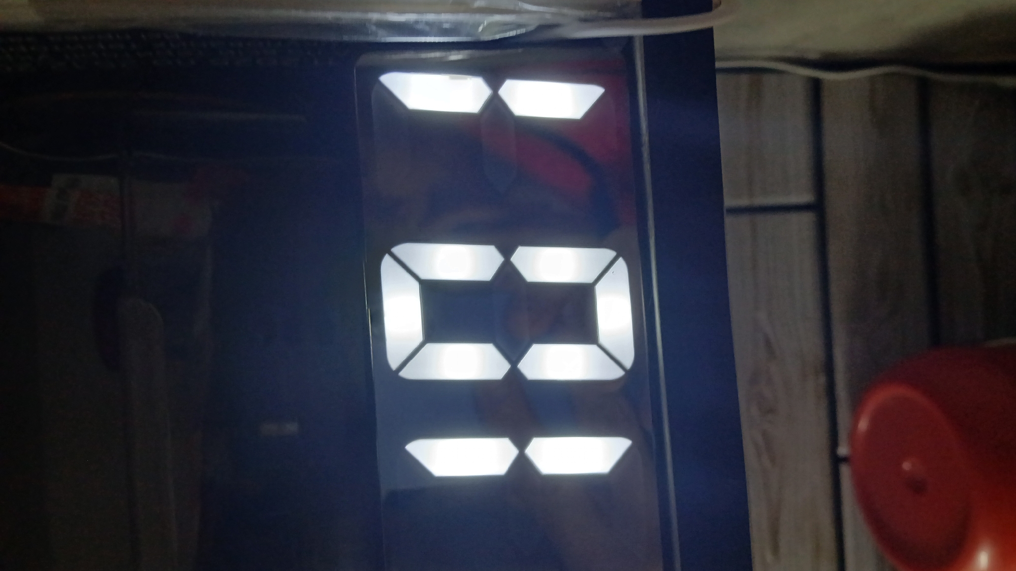 The image shows a digital display illuminated with the number "03". The display is dark and likely part of an electronic device, possibly a clock or timer. The background is dark and indistinct. A white power cable is visible in the upper left corner. Adjacent to the right side of the digital display is a vertical background of white wooden planks or paneling. A red object is barely visible in the lower right corner of the image.
Location: Unknown, Unknown.