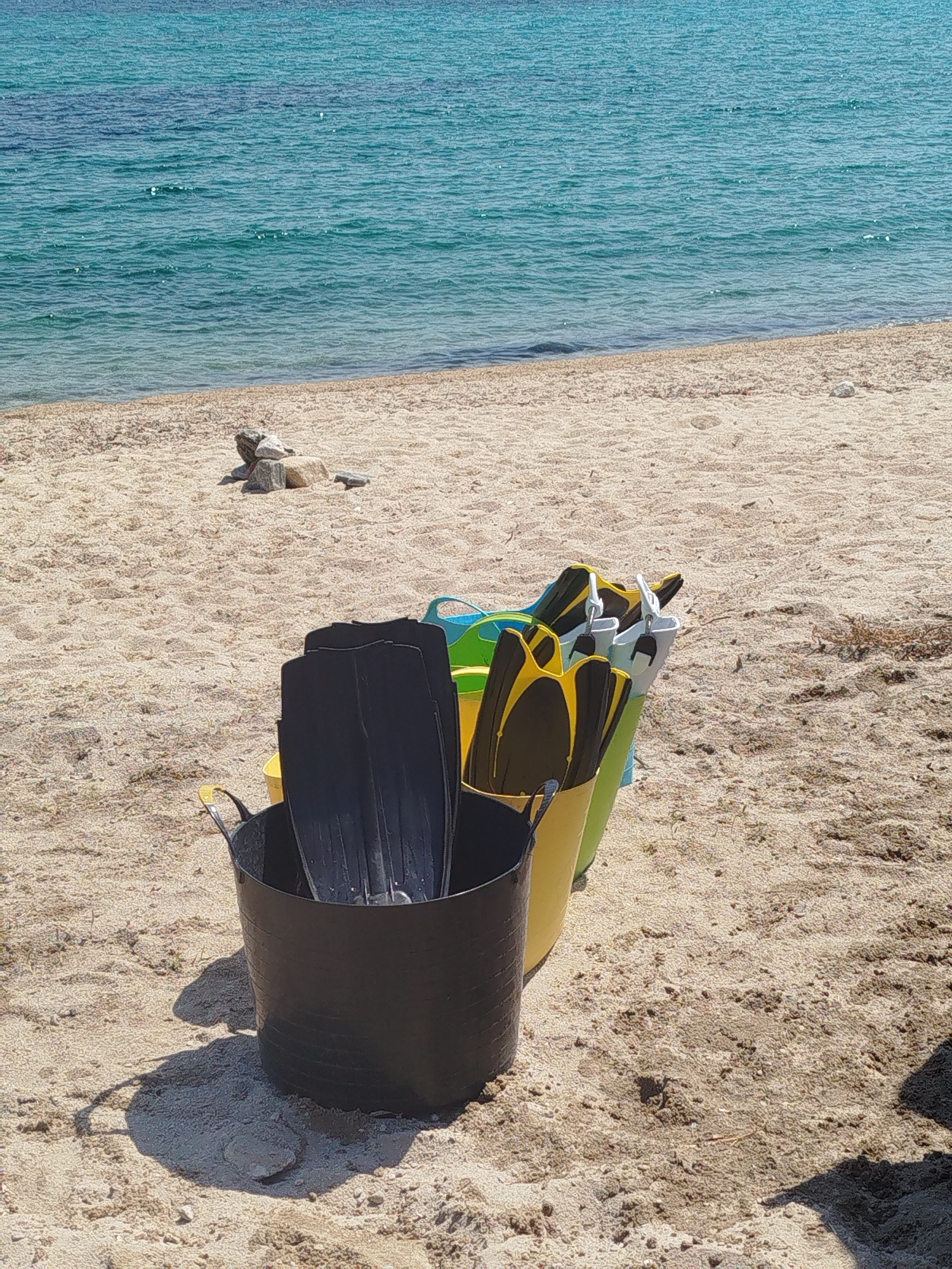 The image depicts a sunny beach scene on a sandy shore with the turquoise sea in the background. In the foreground, a black bucket contains several black swim fins, and behind it, two more buckets, one yellow and one green, are visible, also holding swim fins, some of which are yellow and black. The sand is textured with small pebbles and debris. A small pile of rocks is present further up the beach, near the water's edge. The scene suggests preparation for water activities. The location is identified as Sithonia Municipal Unit, Greece.