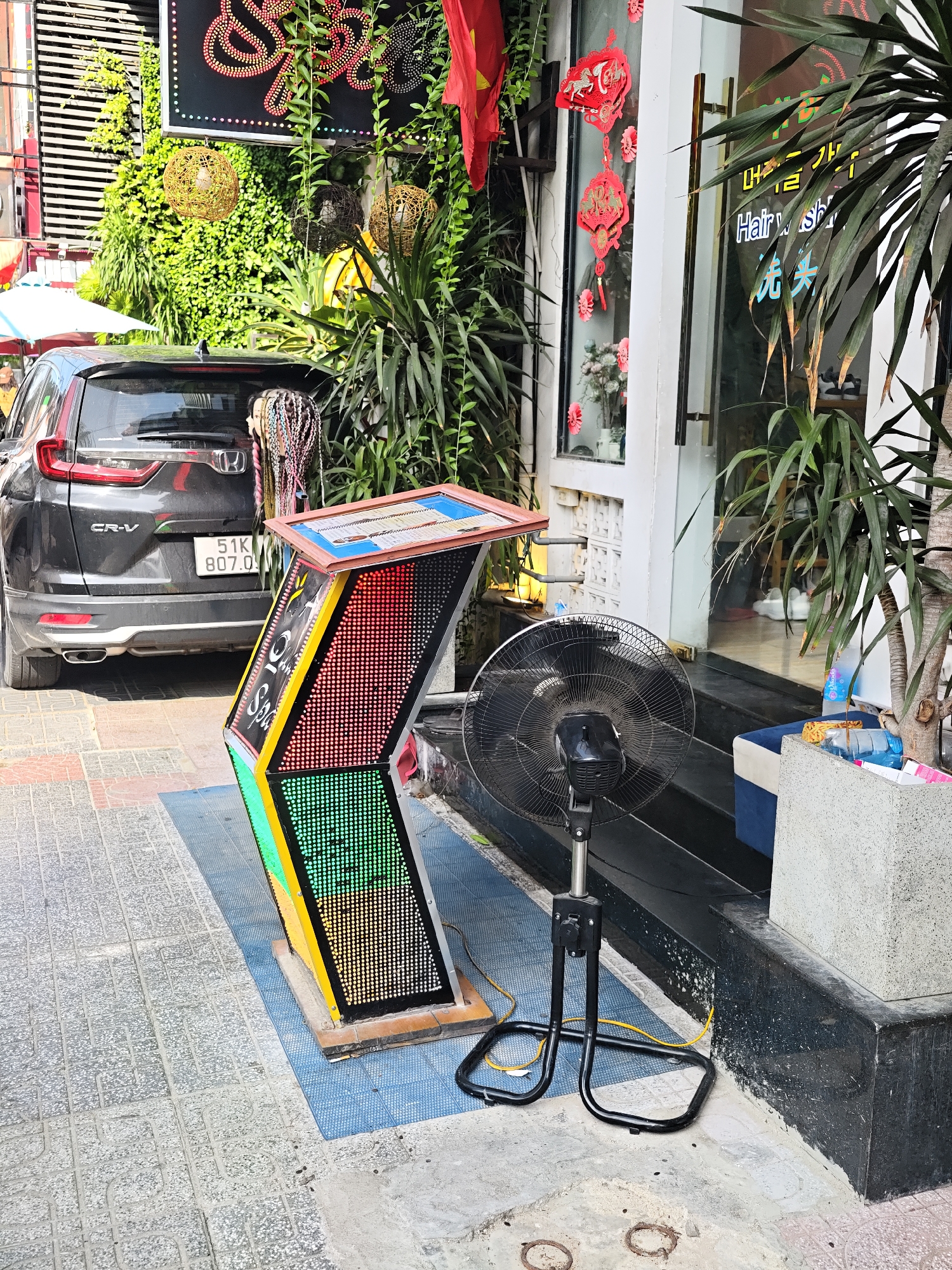 The image depicts a street-level view in Nha Trang Ward, Vietnam, on a sunny day. In the foreground, a dark grey Honda CR-V is parked on the left, with its rear visible. To the right of the car is a tall, colorful, angular electronic display board with a screen composed of small, illuminated dots in red, orange, yellow, and green. Above the display board, a black oscillating fan with a metal grille stands on a black metal tripod base, its power cord snaking across the ground.
Behind the car and display board, lush green foliage cascades down, suggesting a densely vegetated area. A portion of a building with a glass storefront is visible on the right, with the words "Hair Wash" and what appear to be Vietnamese characters on the glass. Red decorative lanterns and fabric banners hang from the building, adding to the festive atmosphere. The ground is paved with a mix of grey patterned tiles and concrete. The overall scene suggests a commercial area with a vibrant, possibly tourist-oriented, environment.