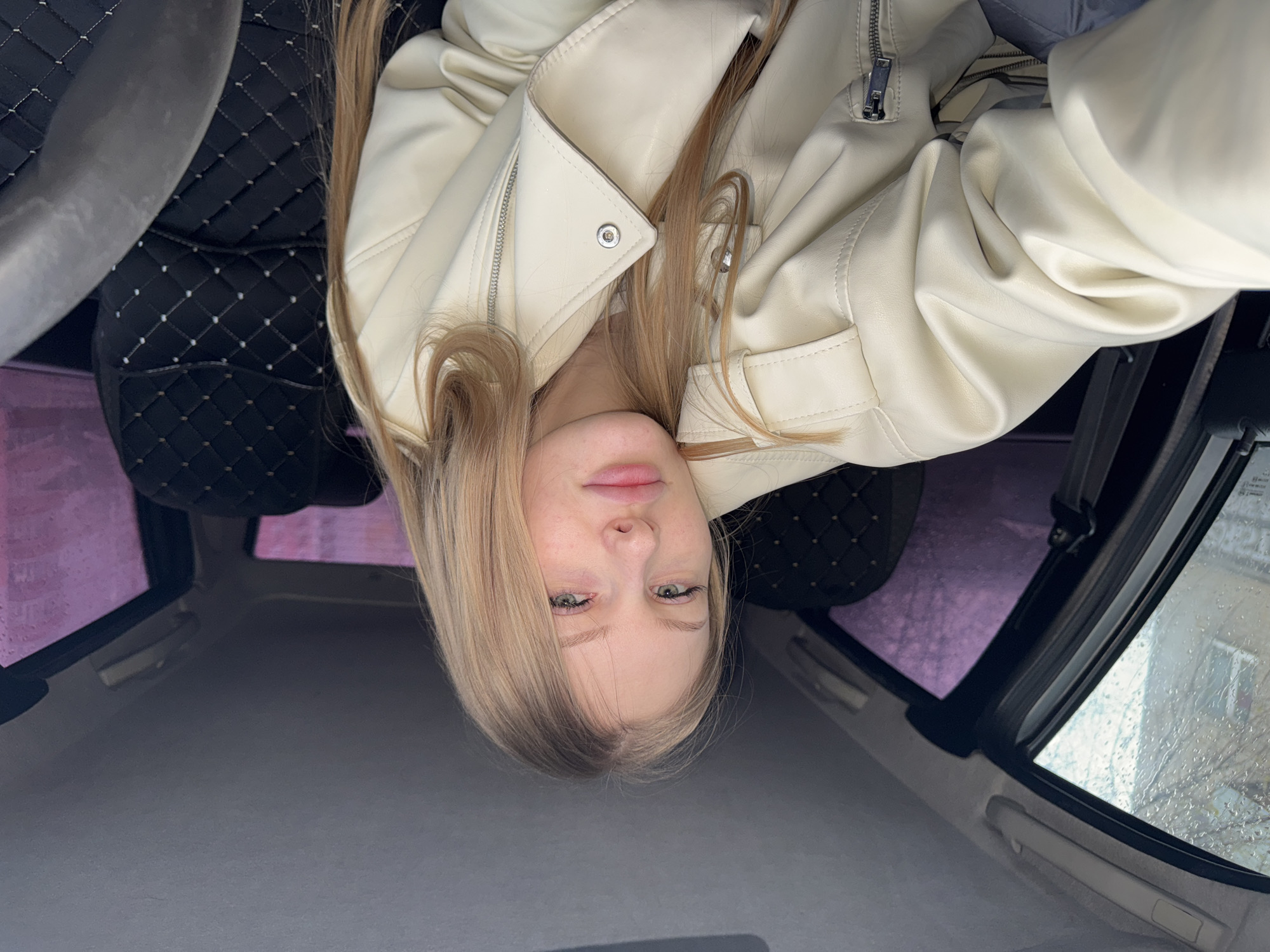 A young woman with blonde hair and light eyes is sitting inside a car, seemingly in Perm, Russia, as suggested by the visible Russian text on the car window. She is wearing a light-colored leather jacket. The car interior features black seats with a diamond-patterned design. The weather outside appears rainy, with raindrops visible on the window. The buildings outside are blurred and indistinct, indicating the photograph was taken from inside the car. The image is oriented upside down.