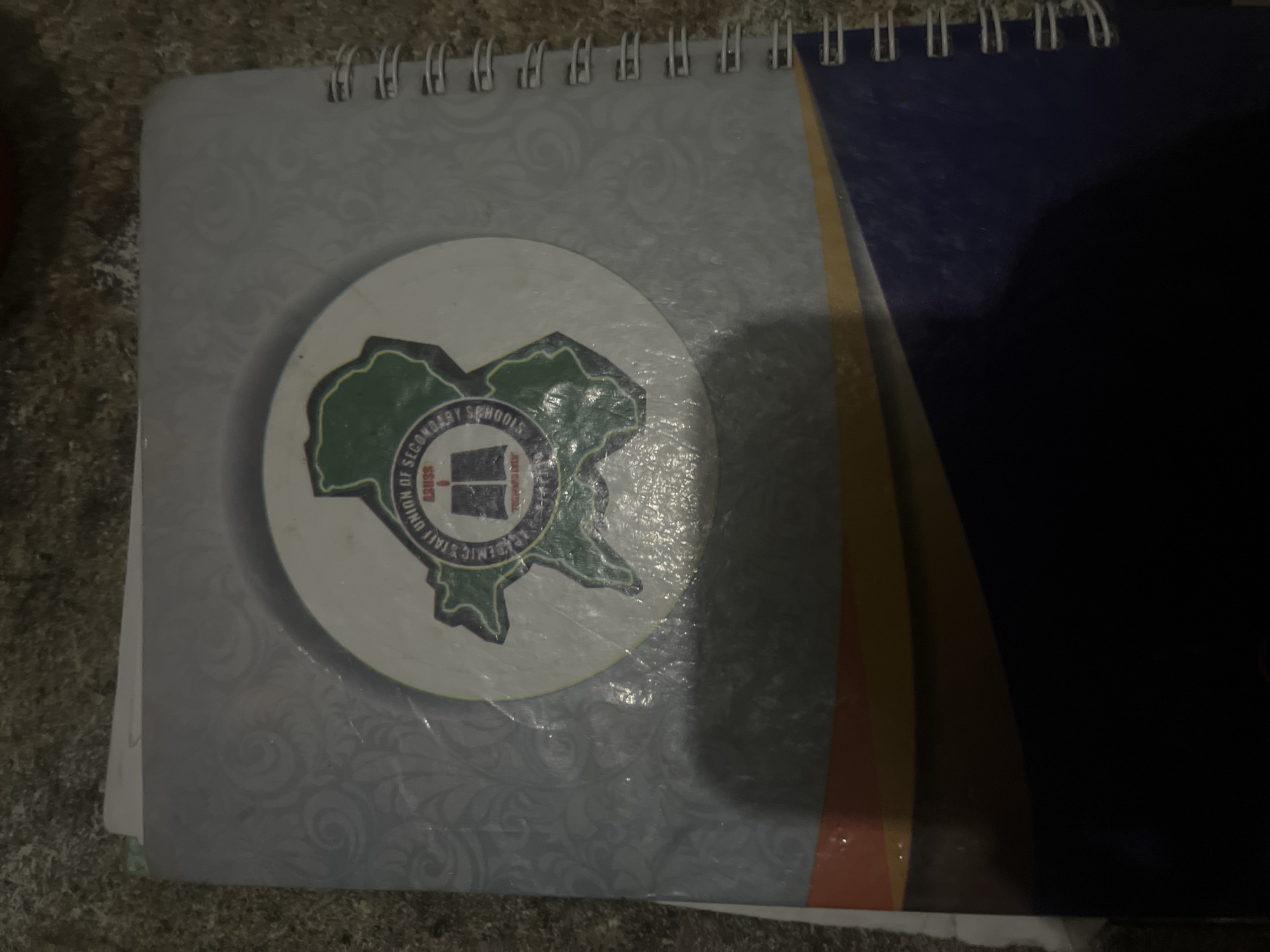 The image shows a close-up of a spiral-bound notebook, likely in an indoor setting given the lighting and texture of the surface it rests on. The notebook cover is predominantly light gray with a faint, swirling pattern. Centered on the cover is a circular emblem. Inside this emblem, a map of a region, outlined in green, is visible. Text is present around the emblem, with "ACADEMIC STAFF UNION OF SECONDARY SCHOOLS" clearly legible in a circular arrangement. Below this, "ASUSS" is written in red, followed by a depiction of an open book and more text in a smaller font. The bottom portion of the notebook cover transitions to a colorful band with orange, yellow, and blue sections. The overall impression is of a notebook belonging to an educational organization. Without further context or visual cues, it is not possible to determine the specific location within Jalingo, Nigeria.