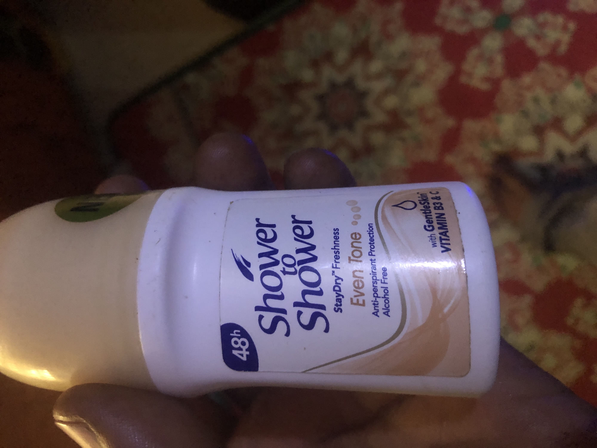 A person's hand is holding a white bottle of "Shower to Shower" roll-on deodorant. The bottle has purple and orange text. The label indicates "48h" protection and "StayDry™ Freshness". Below this, it reads "Even Tone", followed by "Anti-perspirant Protection" and "Alcohol Free". At the bottom, there is text stating "with GentleSkin VITAMIN B3 & C". The background is a blurry, colorful patterned fabric. The image was taken in Kabong, Nigeria.