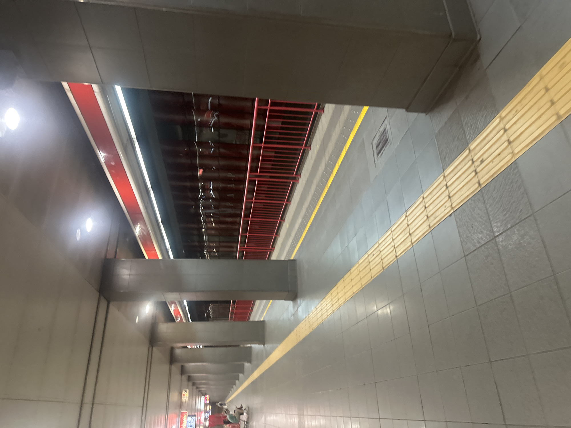 A subway platform in Sofia, Bulgaria, is depicted in the image. The platform is empty except for two individuals sitting on a bench in the distance to the left. The floor is tiled with gray squares, and a yellow tactile paving strip runs along the edge of the platform, indicating the safe zone for visually impaired passengers. A red metal railing separates the platform from the tracks. Large gray concrete columns support the ceiling, which has recessed lighting. The wall behind the railing is a dark color with vertical red accents and reflective surfaces that appear to be part of a train or a tunnel lining. The overall scene conveys a sense of quietness and order on a public transportation hub.