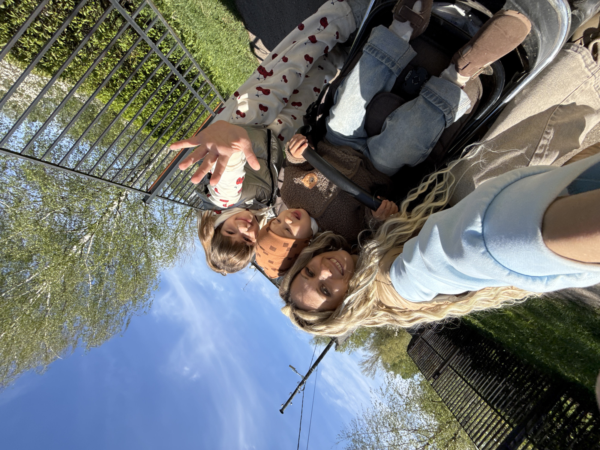A selfie captures a woman and two children in what appears to be a stroller. The woman has blonde hair and is smiling, while the children, dressed in casual clothing, are looking towards the camera. One child is reaching out towards the camera. The location is outdoors, indicated by a blue sky, trees, and a black metal fence. A utility pole is visible in the background. The exact location, Rusanovo, Russia, is based on the user's input and cannot be verified from the image alone.