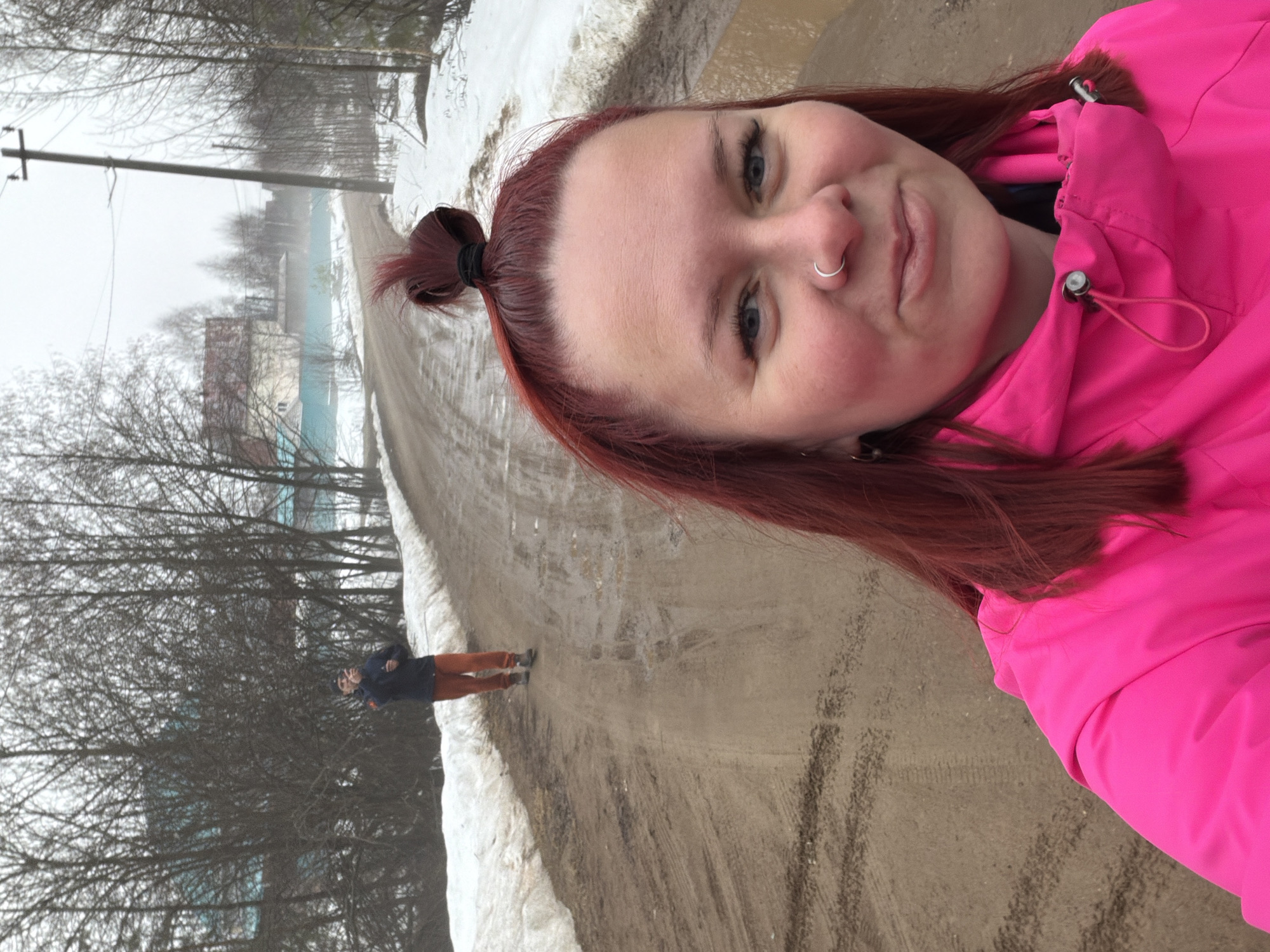 A woman with red hair is taking a selfie on a muddy road. She is wearing a bright pink jacket and has a nose ring. In the background, a man is walking down the road towards a distant building. The environment includes melting snow, trees, and utility poles. The location appears to be in a rural area of the city of Chaikovsky, Russia, based on the landscape and style of the buildings.