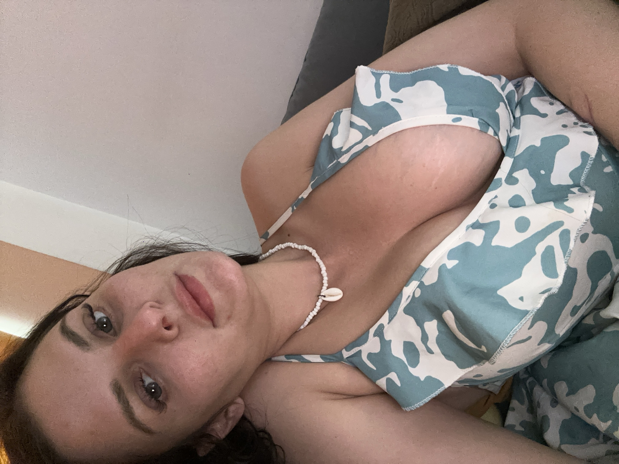 A woman is lying down and looking at the camera. She has dark hair and is wearing a blue and white patterned top. She has a white beaded necklace with a shell pendant. The background appears to be a wall.