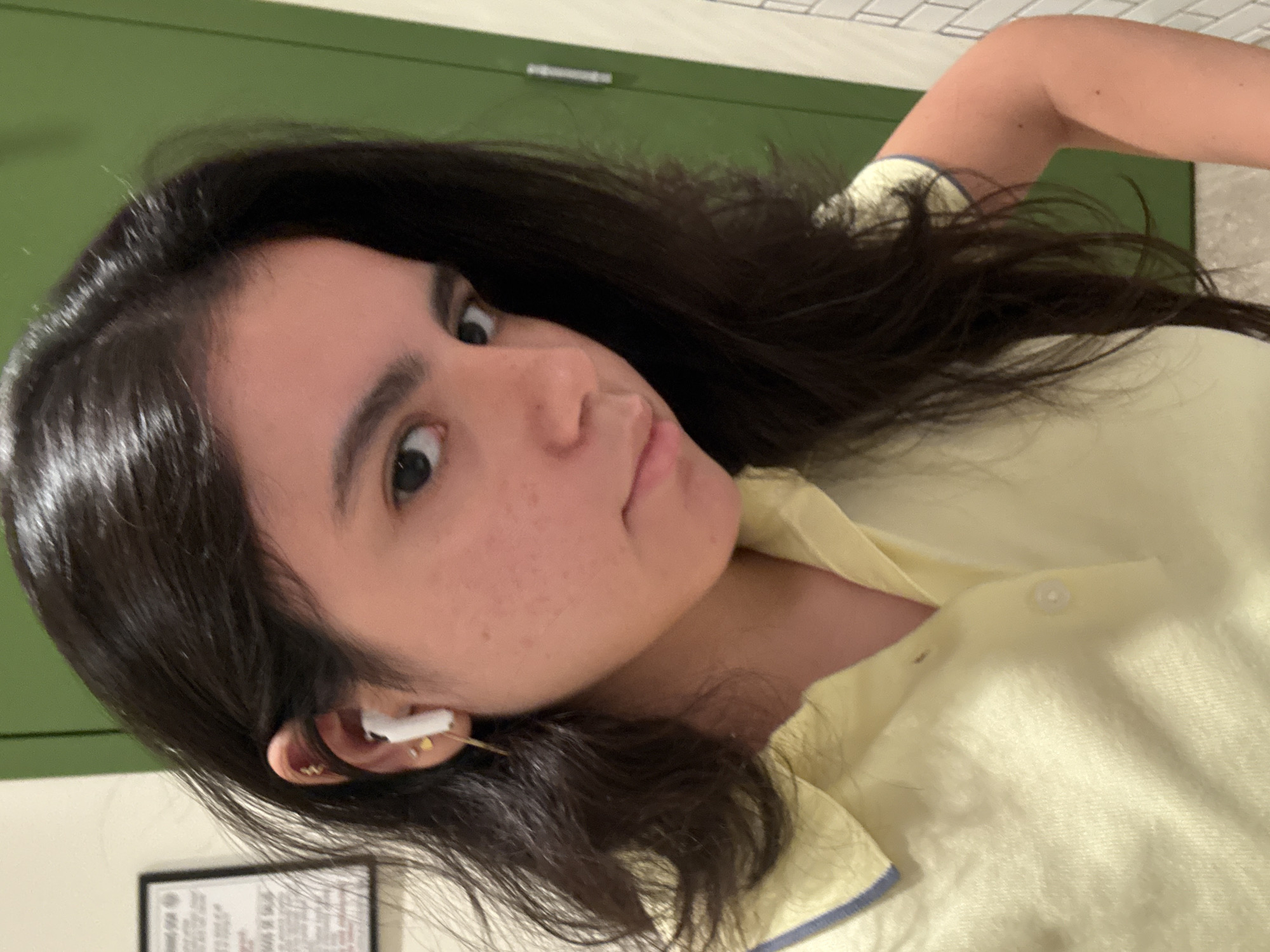 A young woman with dark hair is looking at the camera. She is wearing a yellow shirt with a blue trim on the collar and sleeves. She has a cotton swab in her ear. The background appears to be a room with a green door, white tile, and a framed document hanging on the wall. The location is the city of Austin, United States of America.
