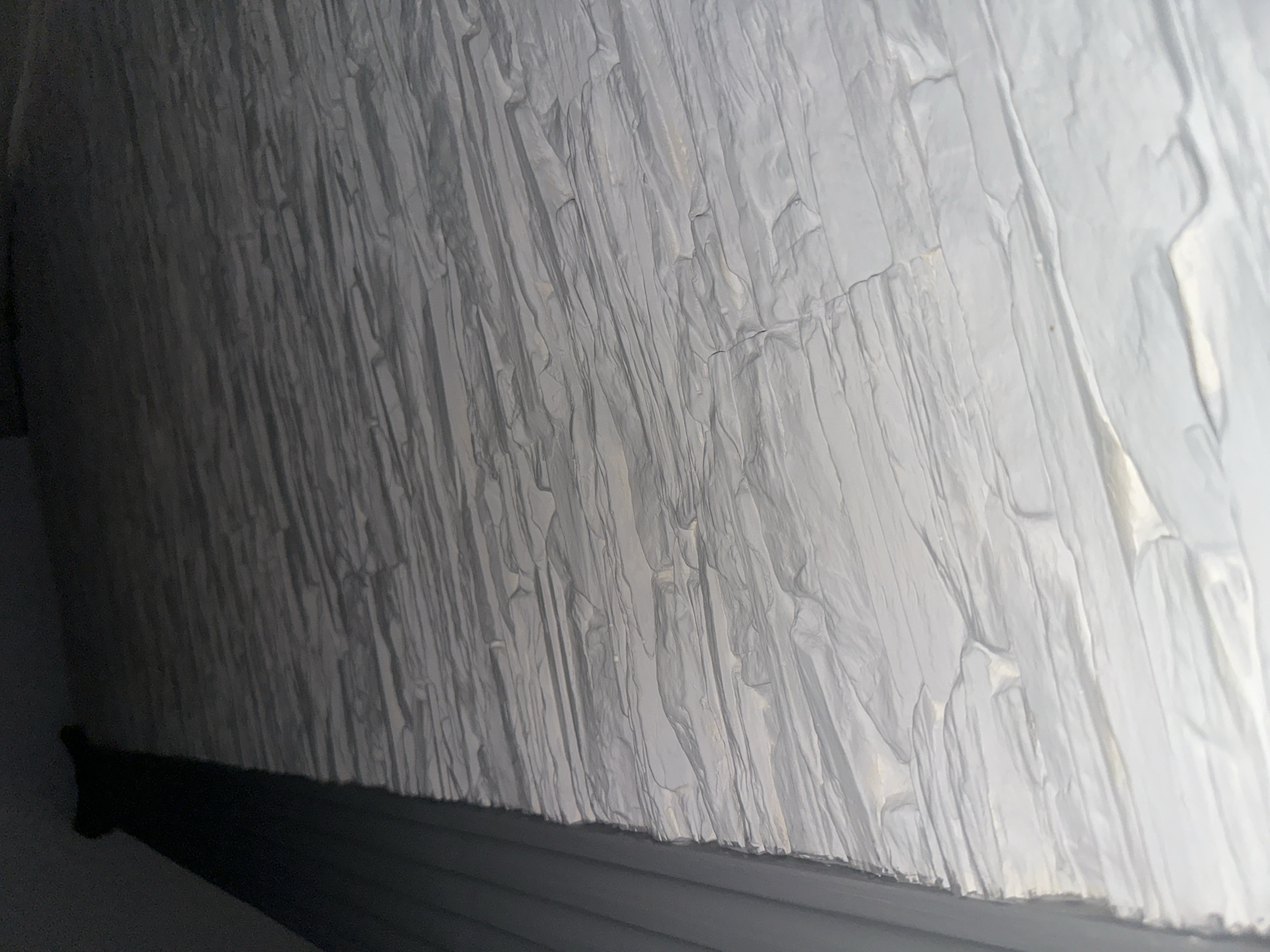 The image shows a close-up view of white, vertically-oriented material with a crumpled or wrinkled texture. The material appears to be a type of fabric or plastic sheet, potentially used as a covering or insulation. A dark, horizontal line is visible at the bottom of the frame, possibly part of a wall or other structural element against which the material is positioned. The location and further context are unknown.