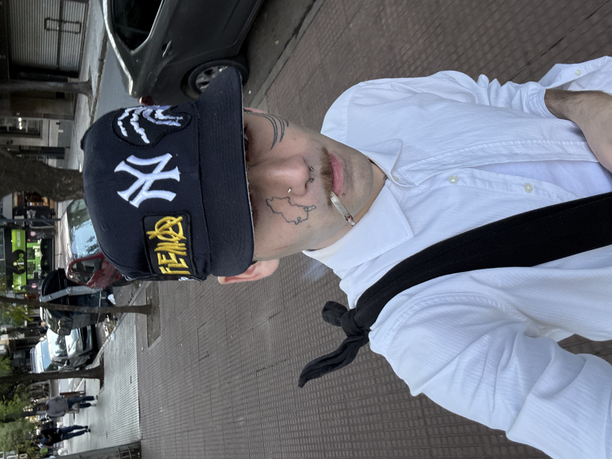 A man stands on a paved sidewalk in Buenos Aires, Argentina. He wears a black baseball cap with white "NY" lettering and yellow detailing, a white button-down shirt, and a black strap or bag across his chest. Tattoos are visible on his face, including one depicting a map-like shape near his nose. A cigarette is held in his mouth. Street scenes with cars, trees, and other pedestrians are visible in the background.