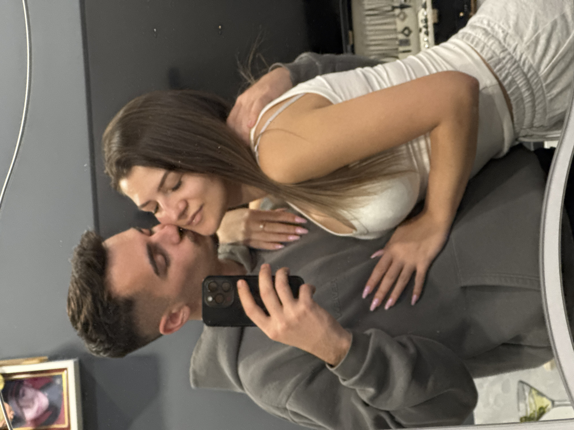A couple is shown in a mirror selfie. The man, wearing a grey hoodie, is holding a black smartphone, taking the photo. The woman, wearing a white top and matching bottoms, has her arm around him and leans in as if kissing his forehead. They are indoors, with items on a shelf visible in the background. The image appears to be taken in a private residence. There is no information available to identify the location in Sofia, Bulgaria.